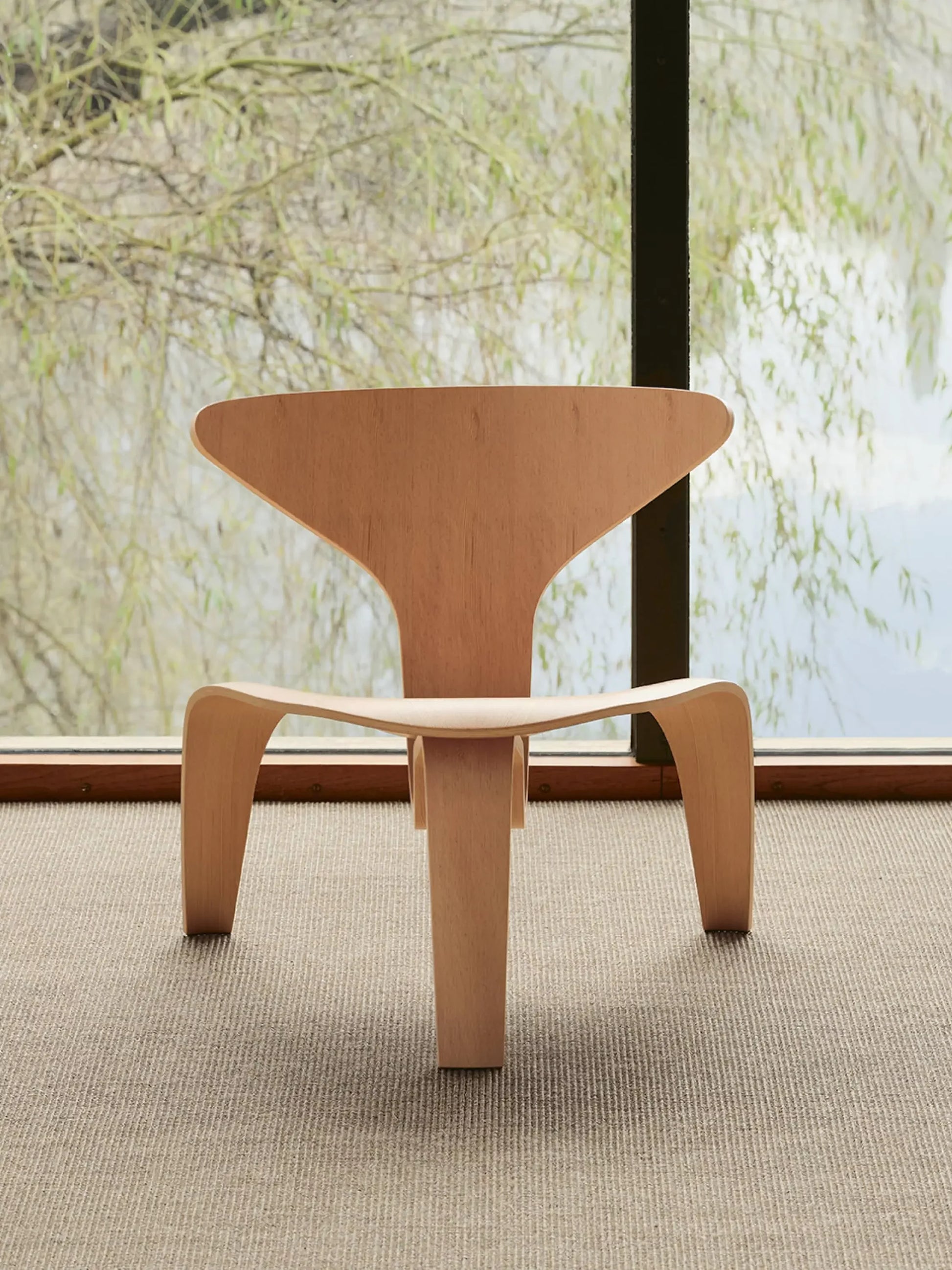 The Fritz Hansen PK0 A Lounge Chair by Fritz Hansen features a triangular backrest and curved legs, placed on a beige carpet before a large window with views of trees and water, highlighting its timeless modern design.