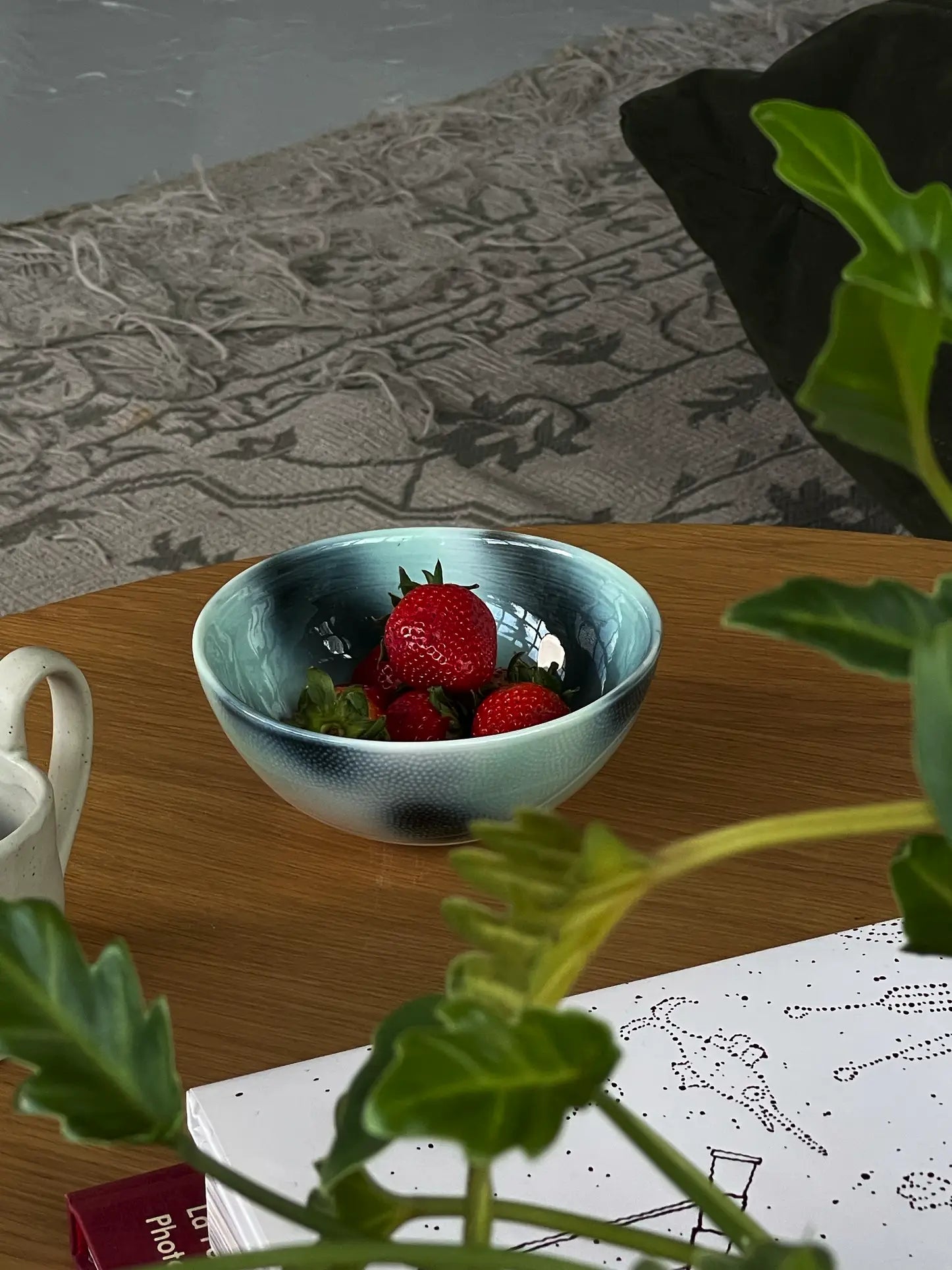 The Frizbee Ceramics BBOL S, a small blue ceramic bowl by Frizbee Ceramics, is filled with strawberries on a wooden table; green leaves, paper, and a patterned rug complete the scene.