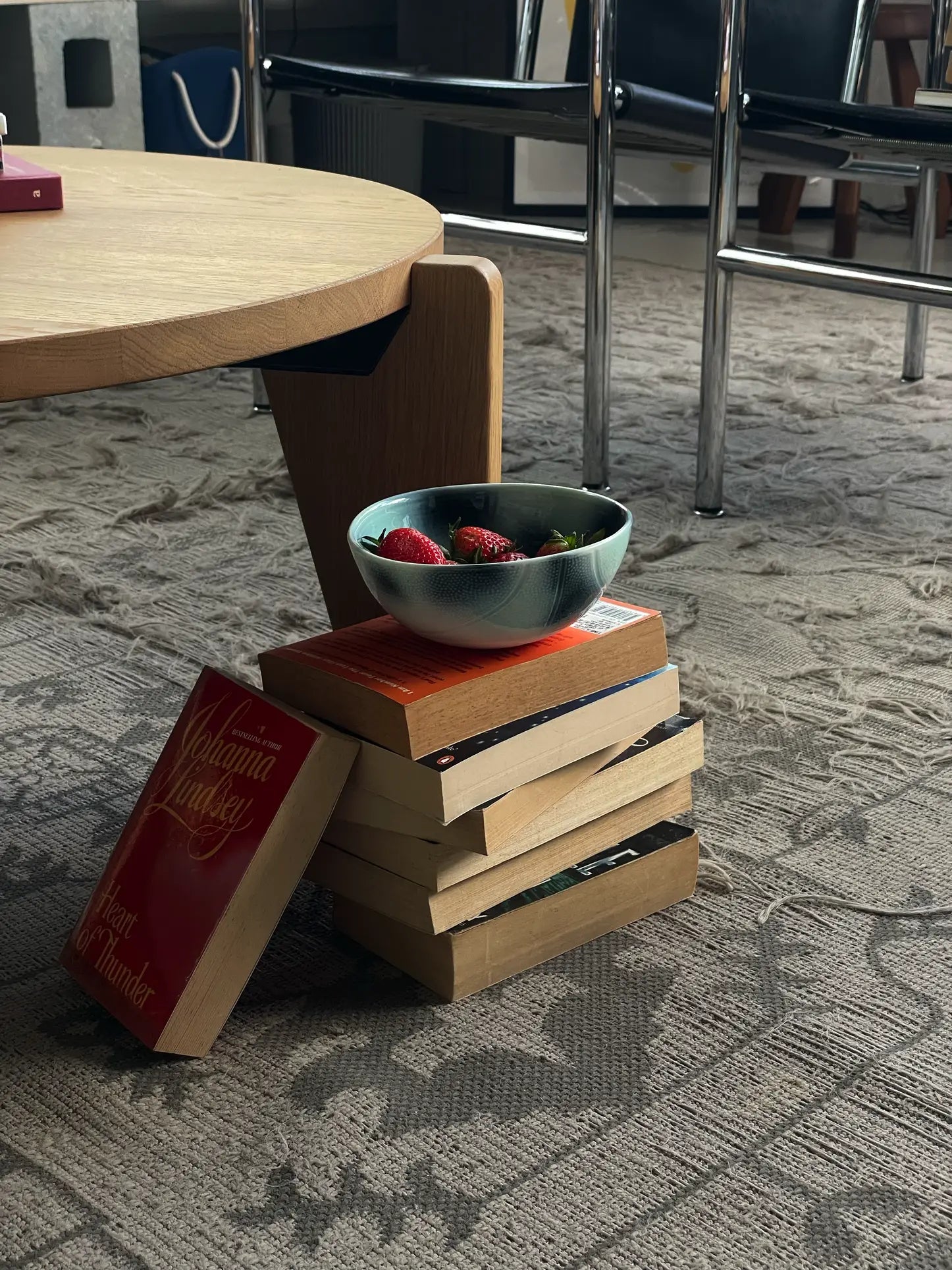 A Frizbee Ceramics BBOL S bowl by Frizbee Ceramics, filled with strawberries, rests on a stack of books by a wooden table atop a patterned carpet, adding artisanal charm to the room with chairs in the background.