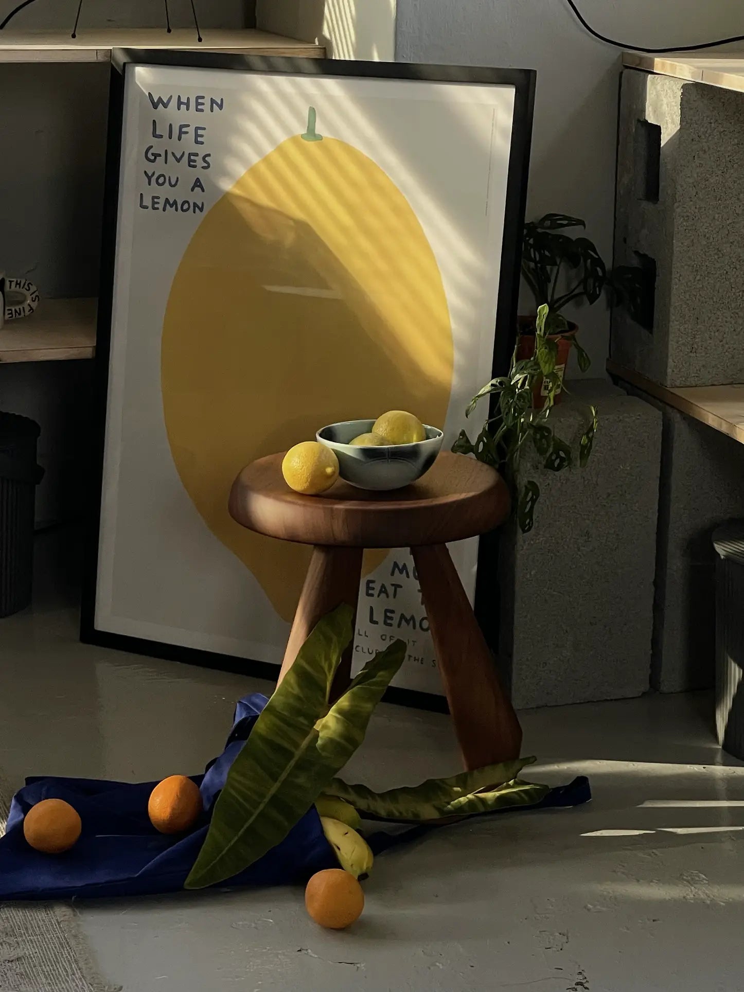 A sunlit room with artisanal charm features a framed lemon poster and, in front, a Frizbee Ceramics BBOL S bowl by Frizbee Ceramics filled with lemons, surrounded by fruit and leaves on a blue cloth.