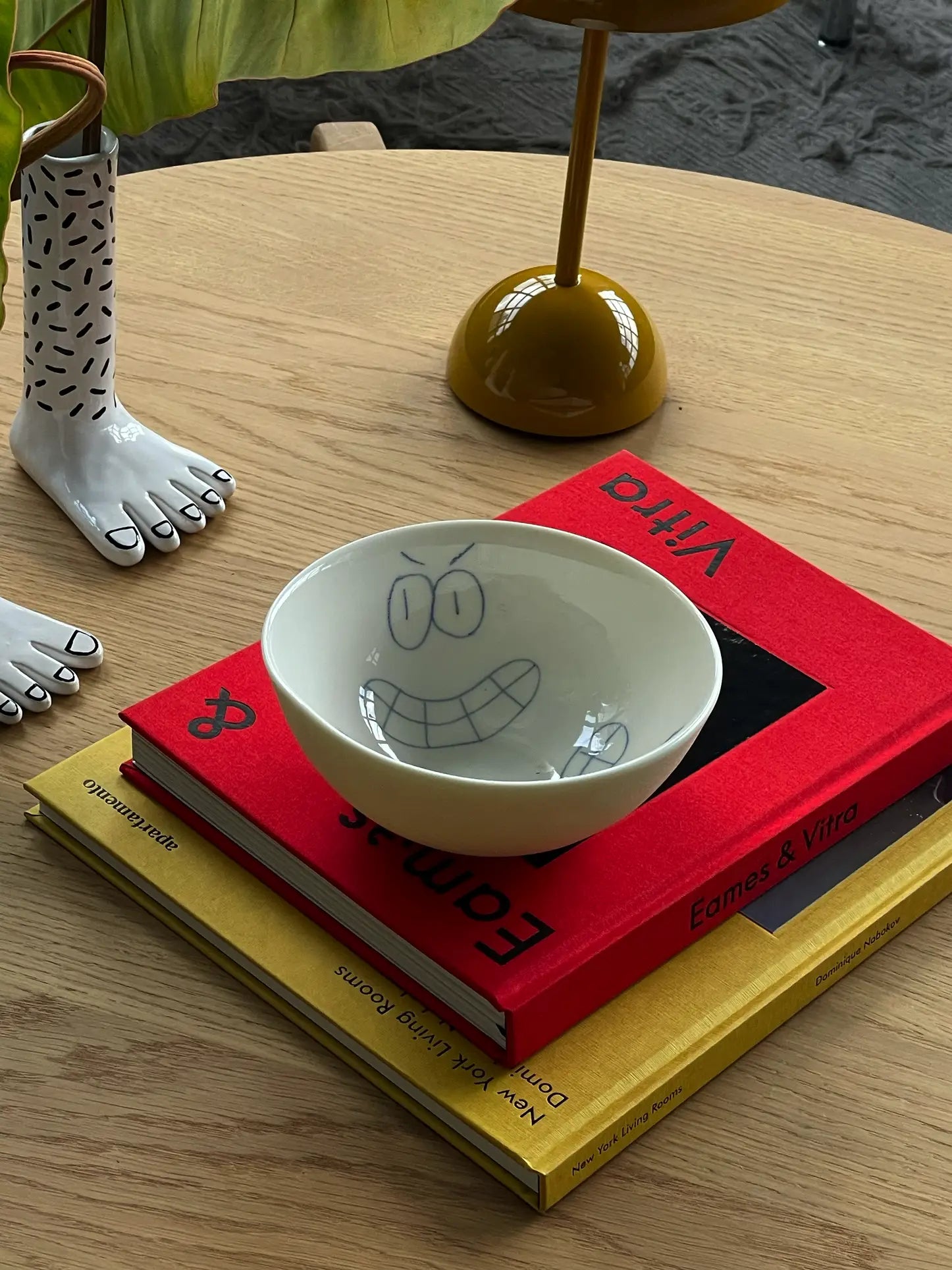 The Frizbee Ceramics BBOL S, a white bowl with a cartoon smile by Frizbee Ceramics, rests on two colorful book designs atop a wooden table beside a sculpted foot decor and a yellow lamp.