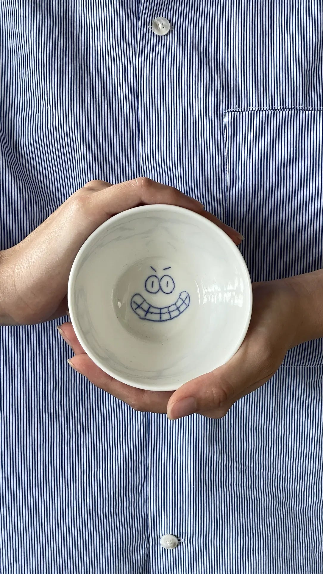 A person in a blue-and-white striped shirt holds the Frizbee Ceramics BBOL S bowl by Frizbee Ceramics, featuring a playful cartoonish smiling face in vivid blue on the inside for an artisanal charm.