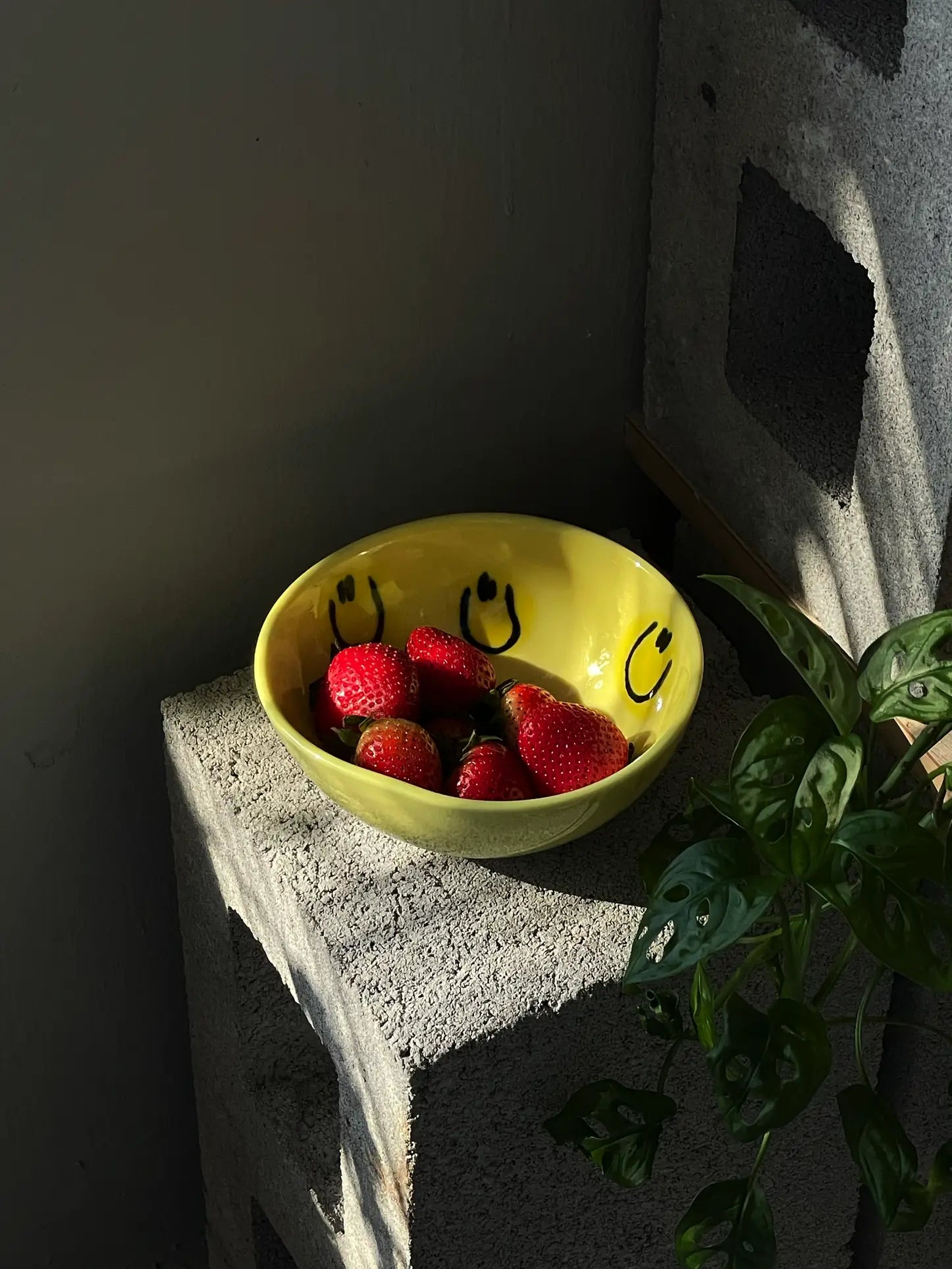 The Frizbee Ceramics BBOL S bowl by Frizbee Ceramics, featuring yellow glaze and black smiley faces, holds strawberries on a concrete block next to a leafy plant, radiating artisanal charm under dappled sunlight.