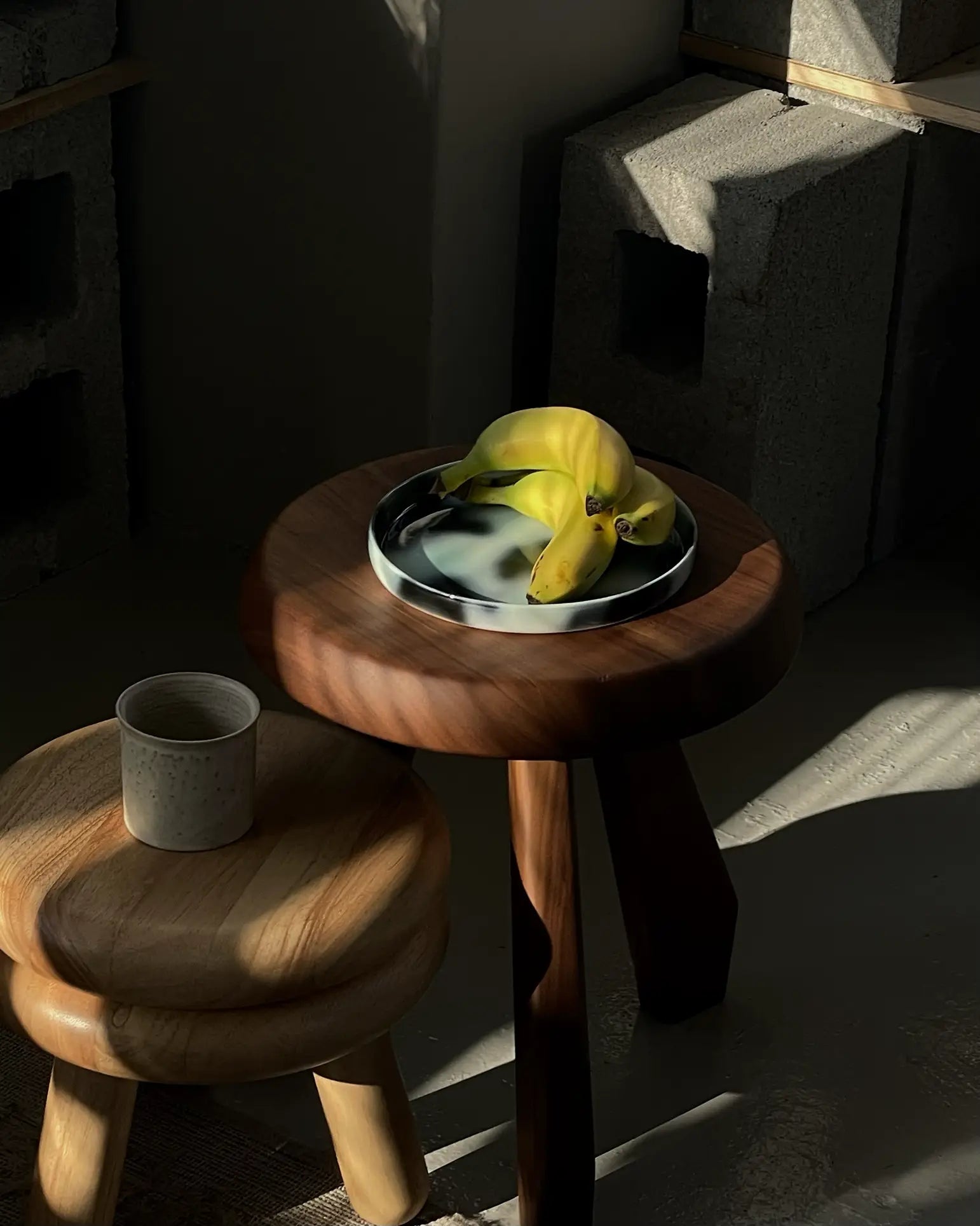 A bunch of bananas sits on the Frizbee Ceramics Baby Plate by Frizbee Ceramics atop a small wooden table. Sunlight and shadows add artisanal charm to the scene, with a ceramic cup nearby and cinder blocks in the background.