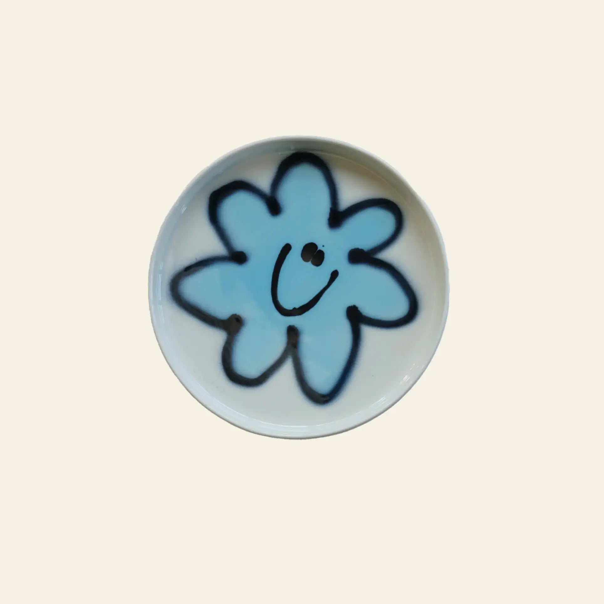 The Frizbee Ceramics Baby Plate by Frizbee Ceramics showcases artisanal charm with a blue flower and black smiley face at the center, set on a simple light background.