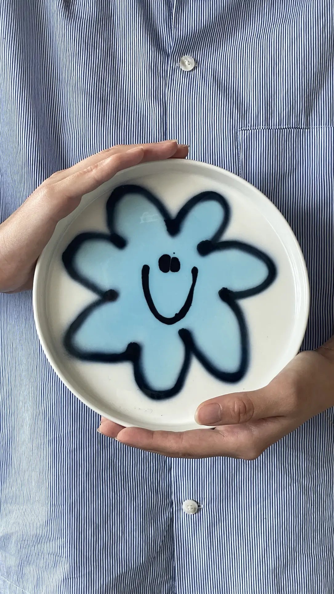 A person in a blue-and-white striped shirt holds the Frizbee Ceramics Baby Plate by Frizbee Ceramics, a round white plate with a blue flower and smiling face at the center, adding artisanal charm to any kitchen collection.
