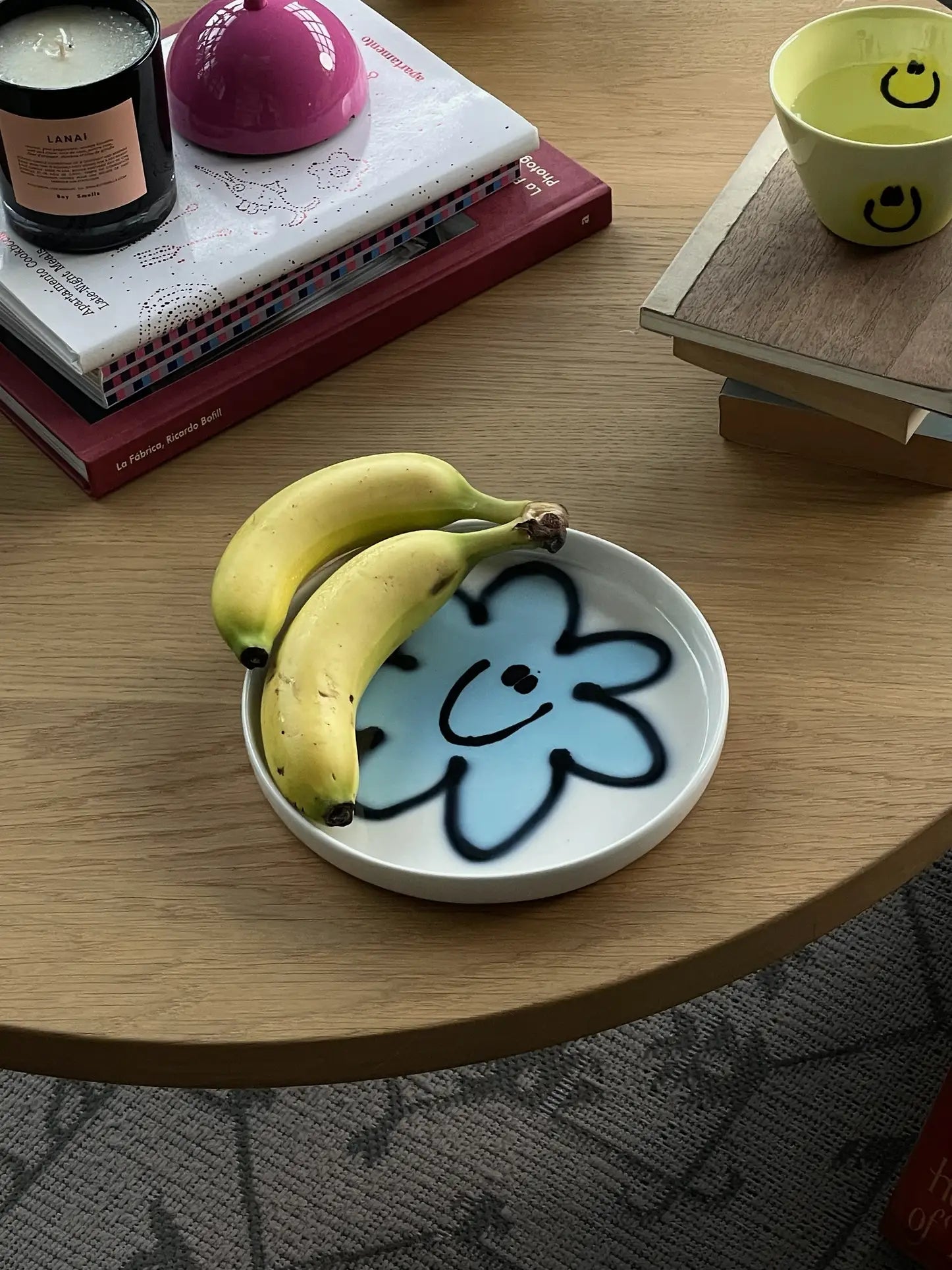 Two bananas rest on a Frizbee Ceramics Baby Plate with a blue smiling flower, bringing artisanal charm to a light wooden table set with books, a candle, and a yellow smiley-face cup.