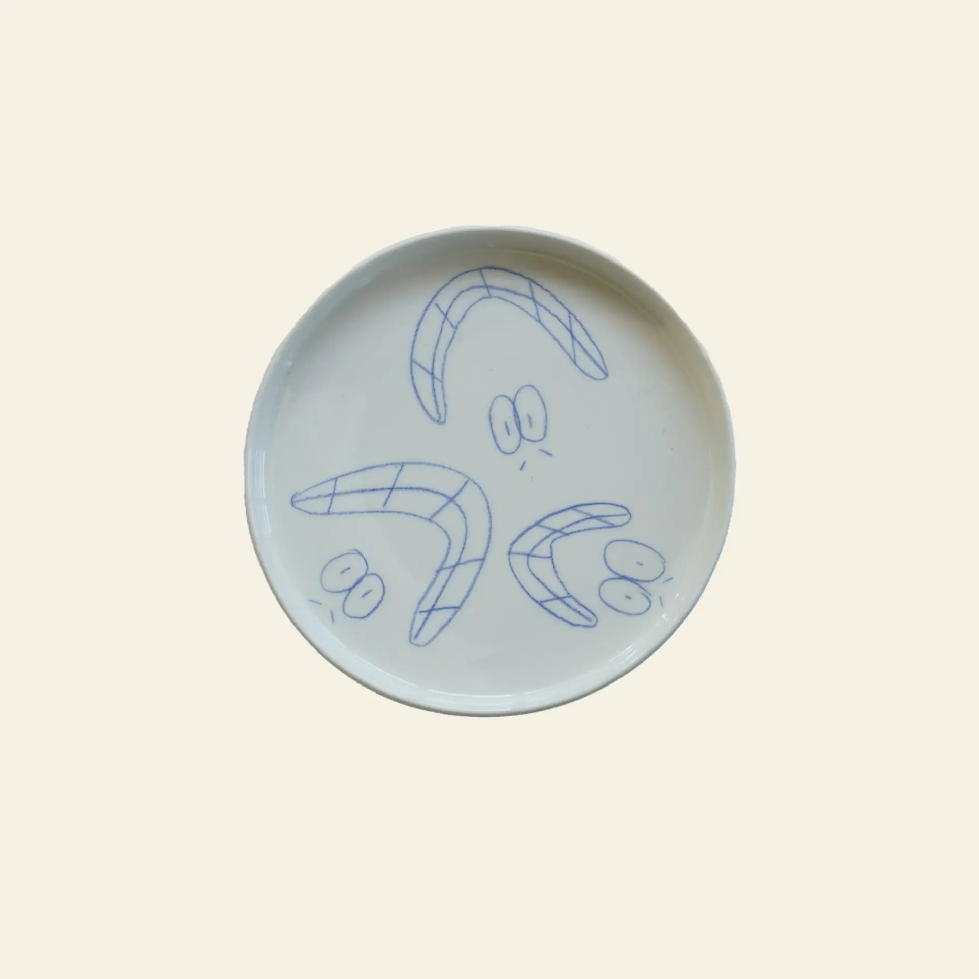 The Frizbee Ceramics Baby Plate by Frizbee Ceramics is a white ceramic plate featuring three blue, boomerang-shaped designs with cartoon eyes, adding artisanal charm to your kitchen collection.
