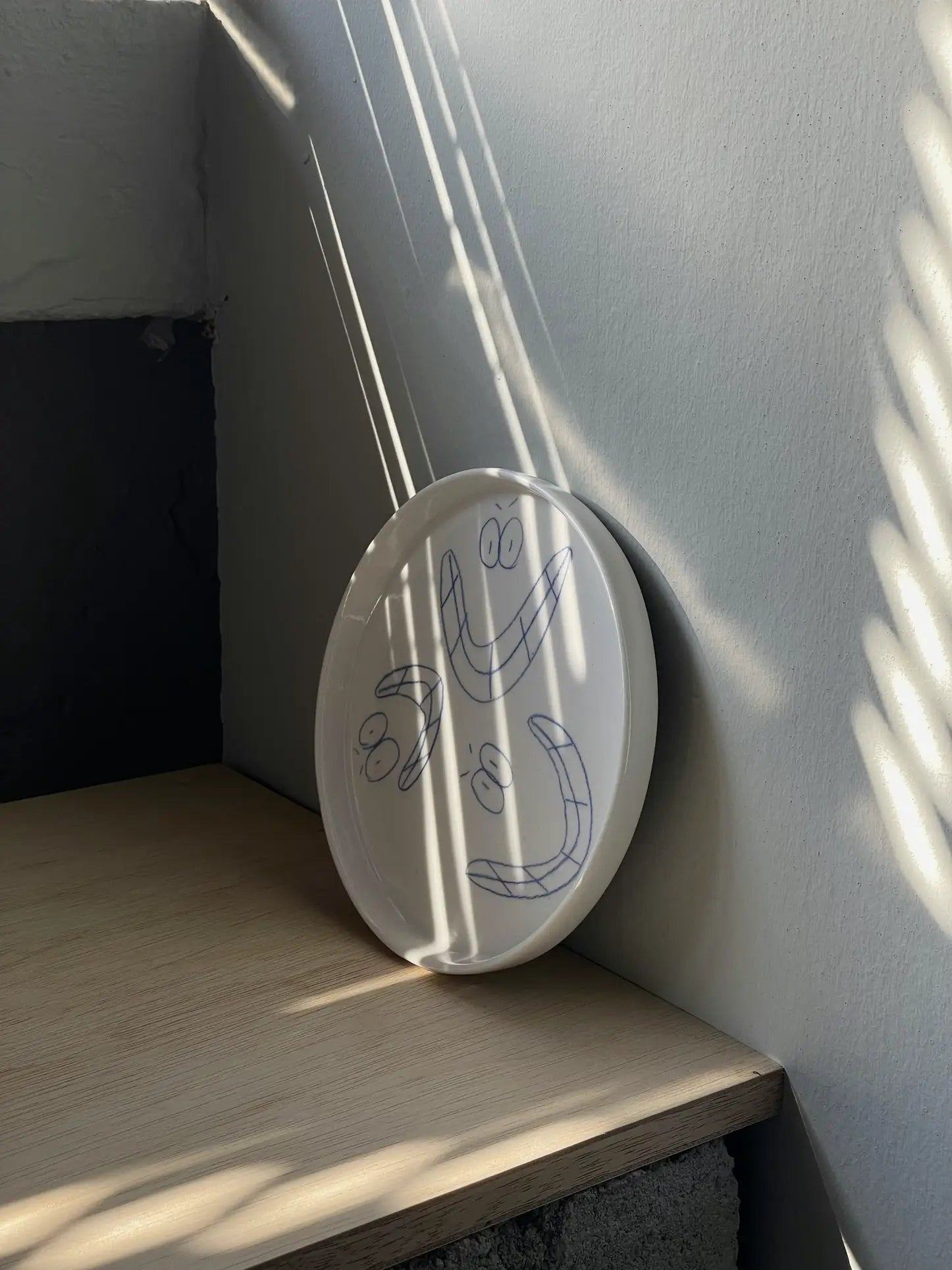 The Frizbee Ceramics Baby Plate by Frizbee Ceramics, featuring three blue cartoon smiling faces, rests against a wall as sunlight plays across its white ceramic surface—a charming and practical kitchen essential.