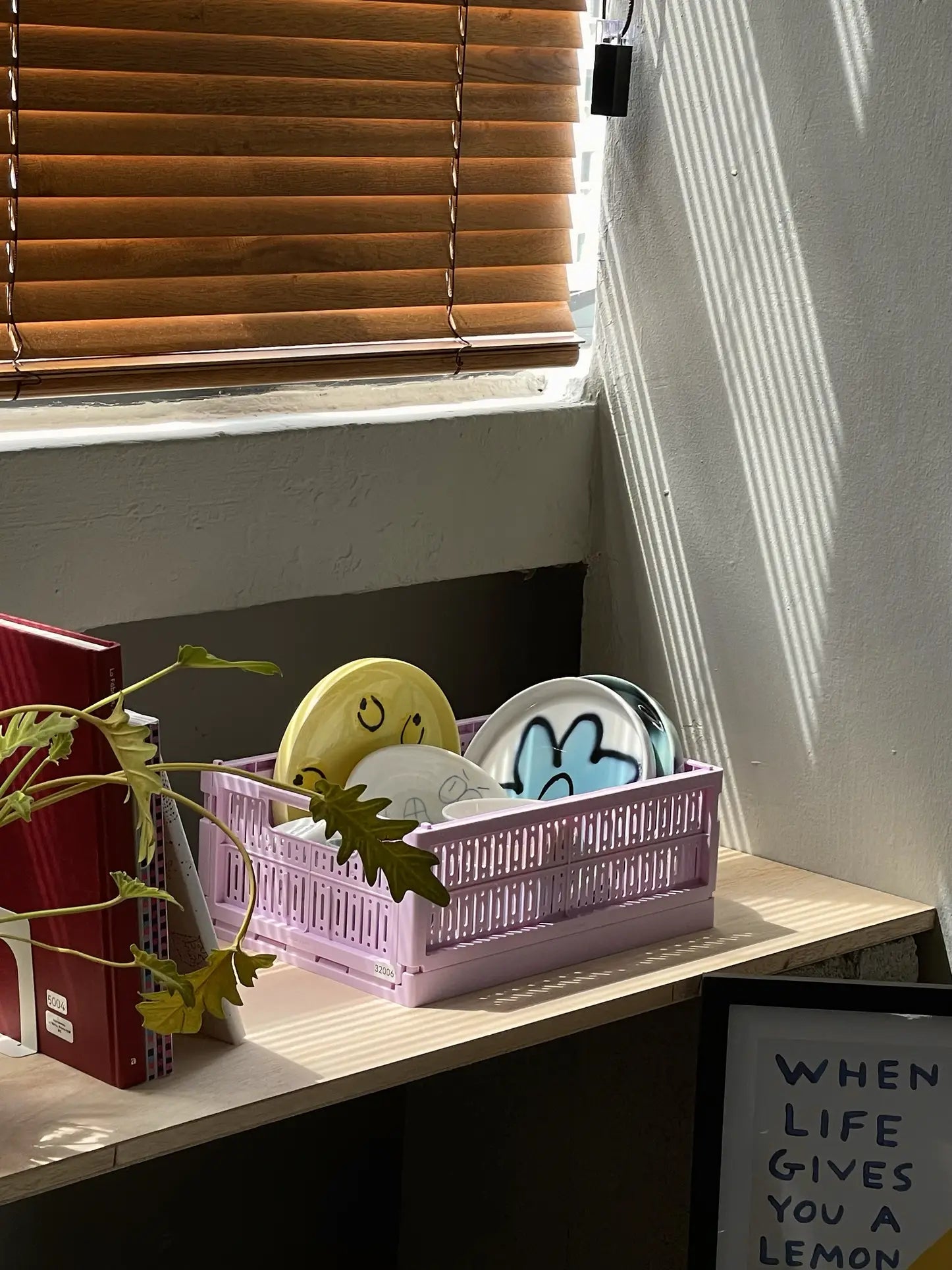 A pink crate holds the Frizbee Ceramics Baby Plate by Frizbee Ceramics, featuring smiley and cloud designs, on a sunlit wooden table near a plant and a framed WHEN LIFE GIVES YOU A LEMON sign, adding artisanal charm.