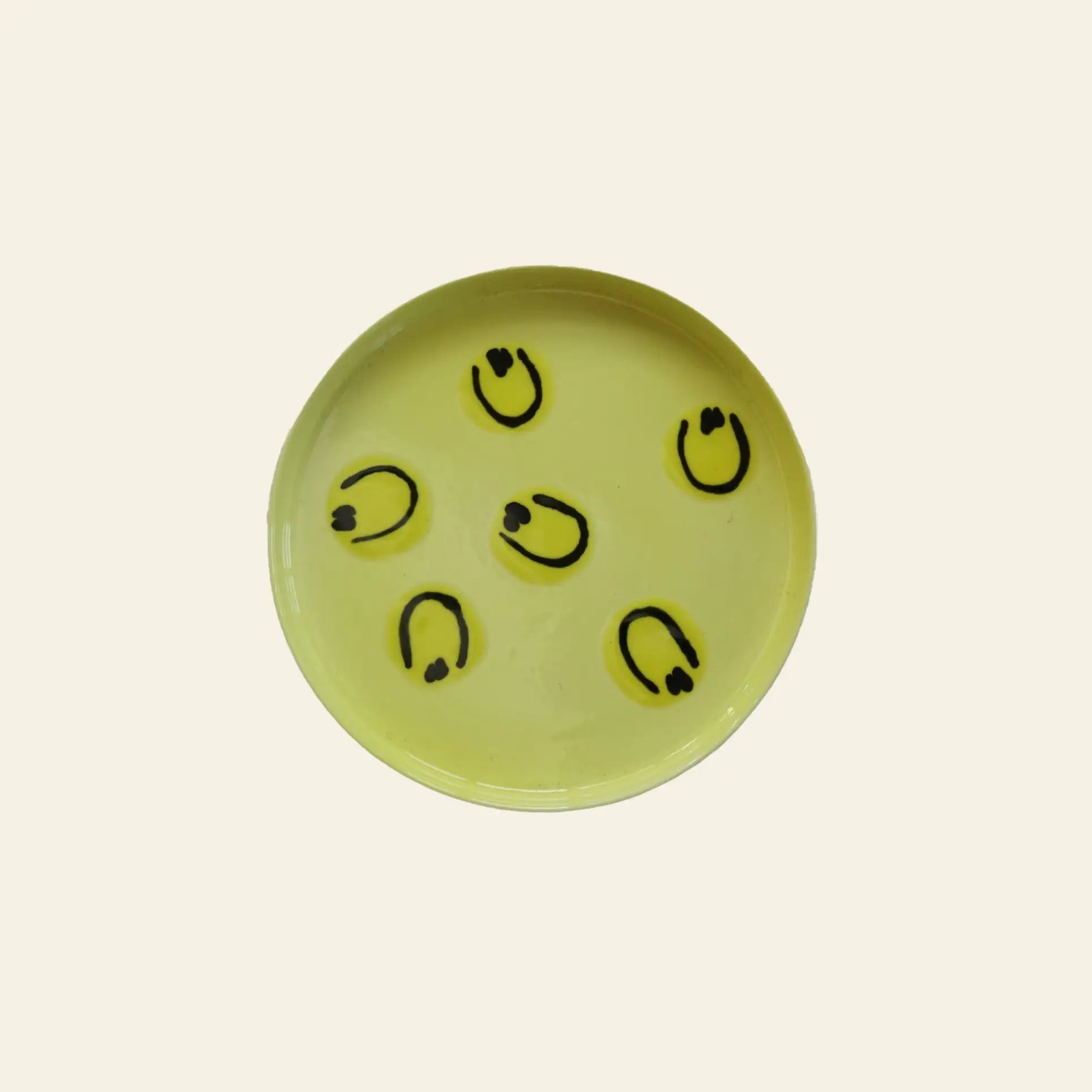 The Frizbee Ceramics Baby Plate by Frizbee Ceramics features six black smiley faces arranged in a circle on yellow, evoking artisanal charm and playful whimsy against a simple background.