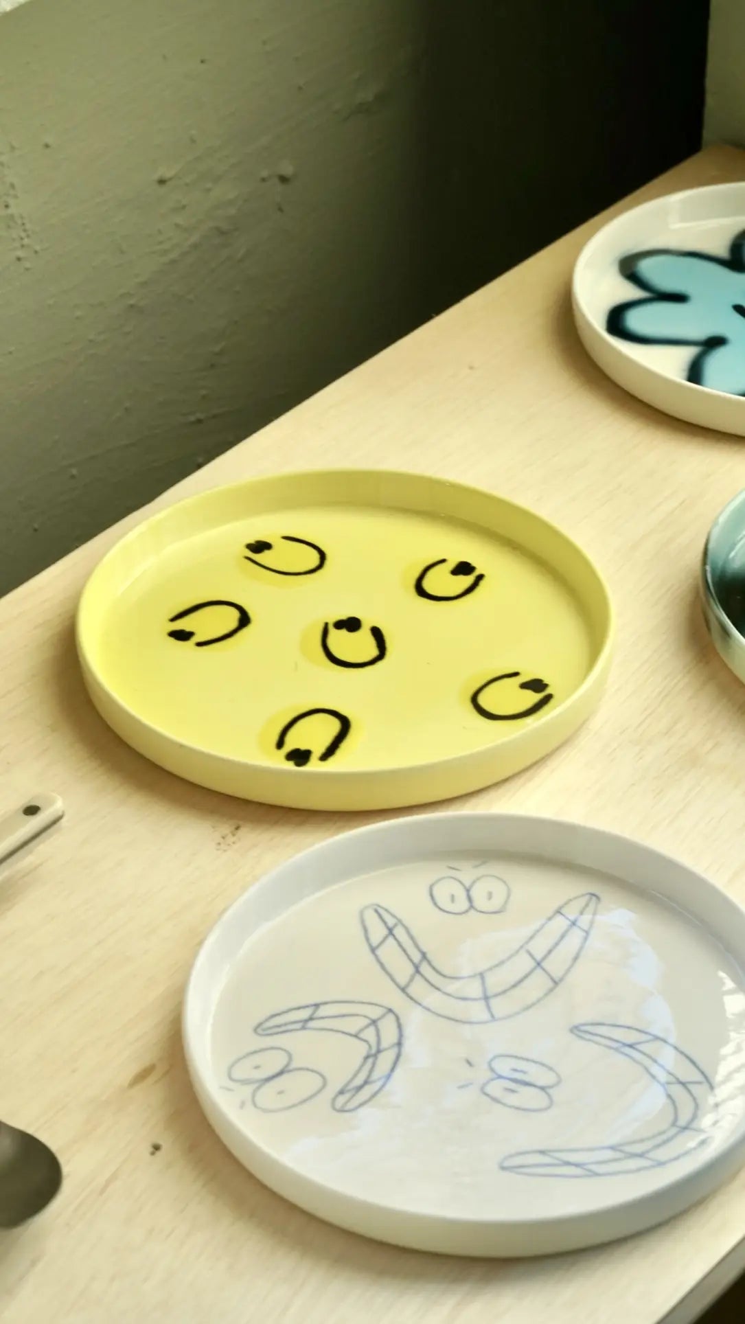 Three decorative plates on a light wooden surface: the Frizbee Ceramics Baby Plate in yellow with a smiling face, a white plate with blue doodle smiles, and a partially visible blue plate—each adding artisanal charm to your kitchen essentials.