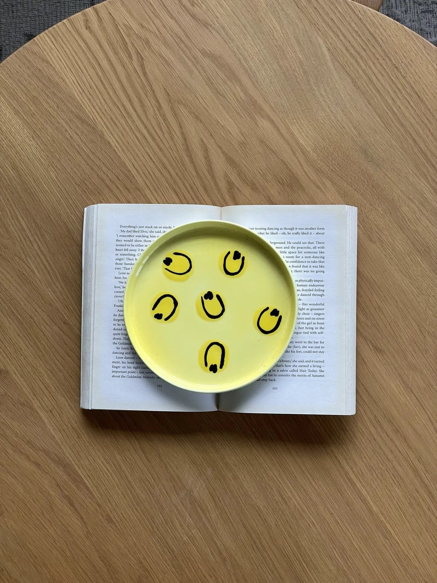 A round yellow Frizbee Ceramics Baby Plate by Frizbee Ceramics, featuring six smiley faces, sits centered on an open book atop a wooden table, partially covering the pages.