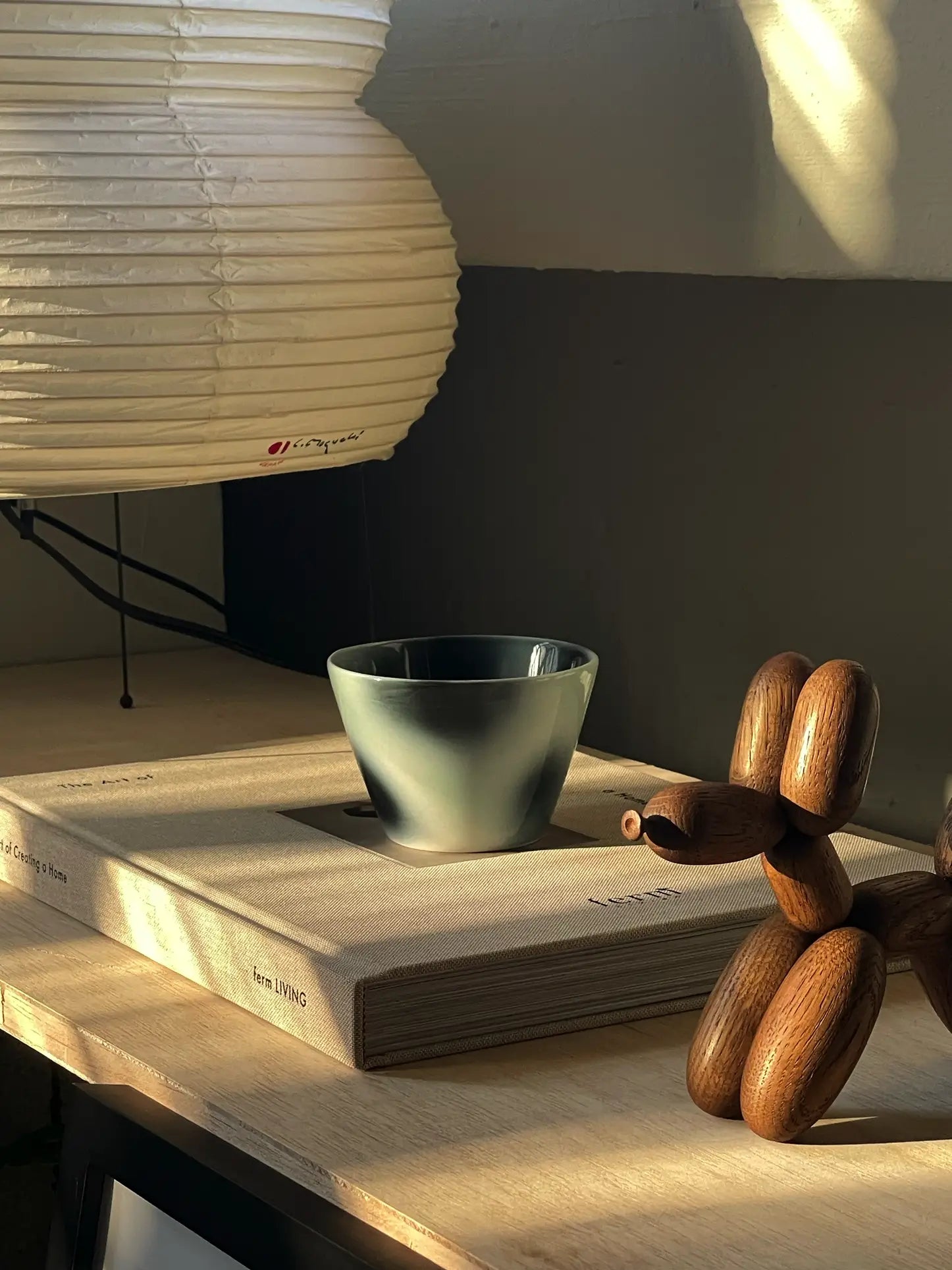 The Frizbee Ceramics Super Cup L by Frizbee Ceramics sits atop a beige book titled Living, next to a sculpted wooden dog and white paper lampshade, all illuminated by warm sunlight on a wooden surface.