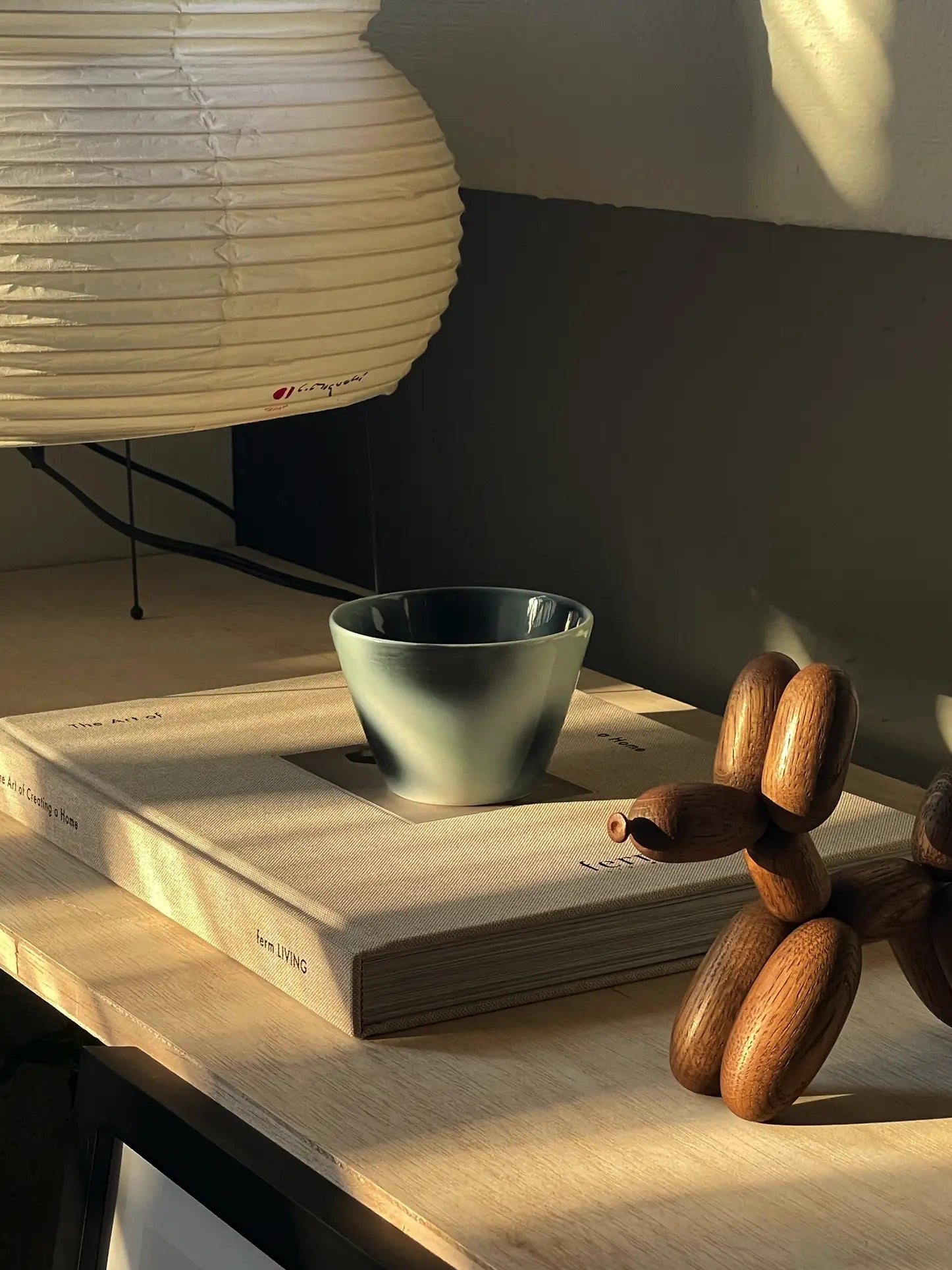 The Frizbee Ceramics Super Cup L by Frizbee Ceramics rests atop a closed book on wood, next to a balloon dog wooden sculpture and a white paper lantern, while sunlight casts gentle shadows.