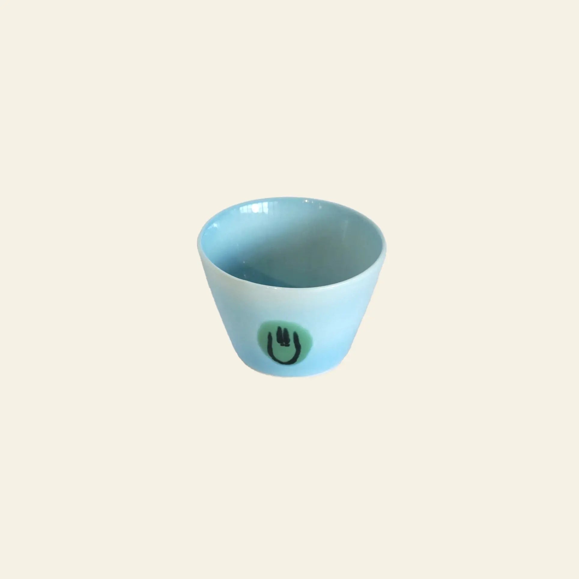 The Frizbee Ceramics Super Cup L by Frizbee Ceramics is a small, light blue ceramic cup with a glossy finish. It features a simple green circle and black smiley face—an artisanal, charming latte companion set against a beige background.