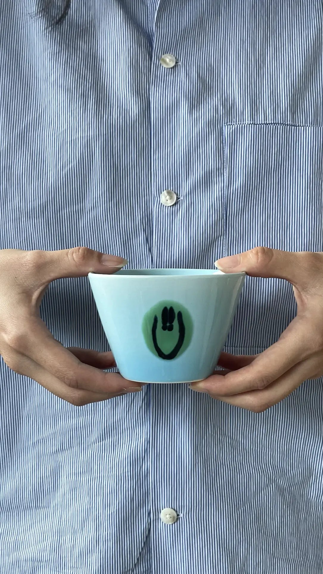 A person in a blue and white striped shirt holds the Frizbee Ceramics Super Cup L by Frizbee Ceramics, a light blue, individually cast bowl with a smiley face design—an artisanal companion perfect for enjoying your latte.