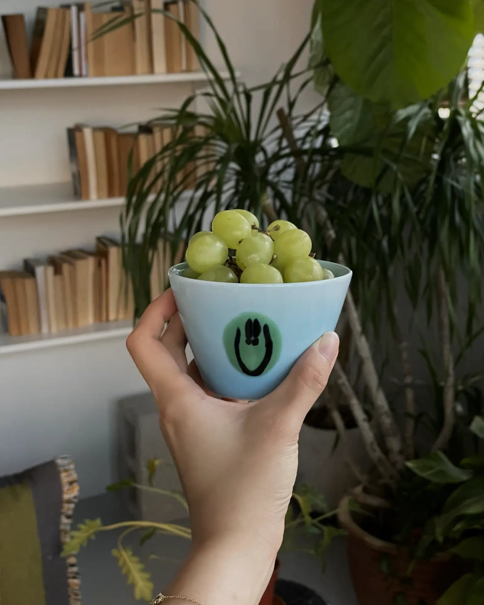 A hand holds the Frizbee Ceramics Super Cup L by Frizbee Ceramics, featuring a smiley face design and filled with green grapes. Its artisanal style pops against bookshelves, vibrant indoor plants, and soft window light.