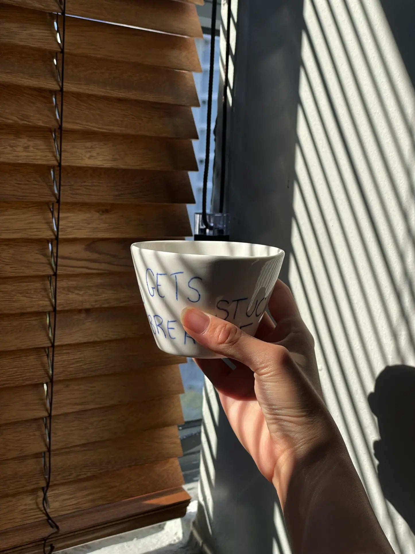 A hand holds the Frizbee Ceramics Super Cup L, a white ceramic bowl with blue script, by a sunlit window with wooden blinds—an artisanal piece from Frizbee Ceramics and an ideal companion for your latte.