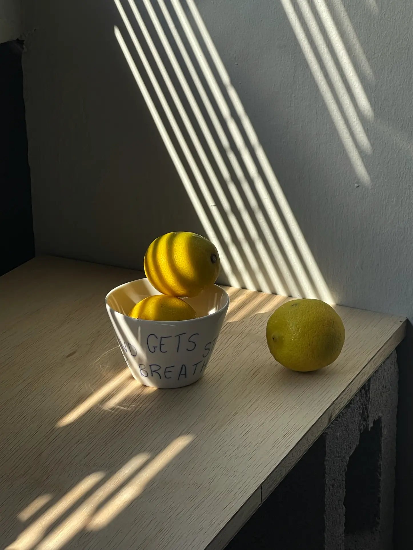 Three lemons rest on a wooden table; two are stacked inside the Frizbee Ceramics Super Cup L, with one beside it. Sunlight casts striped shadows, highlighting the artisanal charm of this inviting Frizbee Ceramics scene.