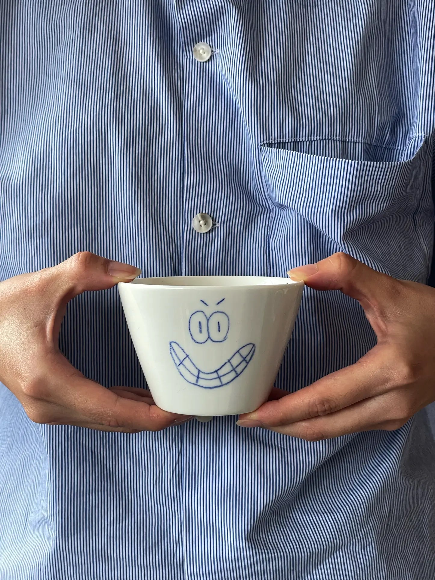 A person in a blue and white striped shirt holds the Frizbee Ceramics Super Cup L by Frizbee Ceramics, a white cup featuring a blue doodle of a smiling face with angry eyebrows—an artisanal charm for your next latte.