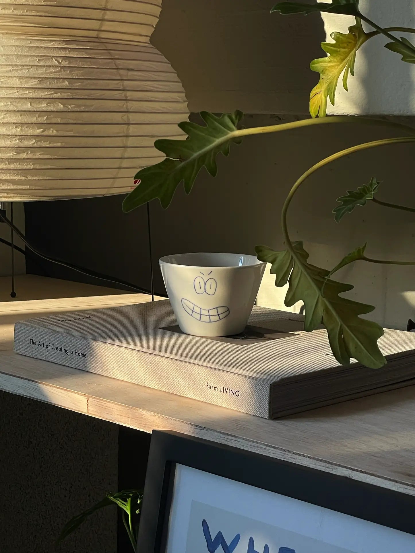 The Frizbee Ceramics Super Cup L, a charming white bowl with a grinning face by Frizbee Ceramics, rests on The Art of Creating a Home, surrounded by sunlight and green leaves for a cozy, inviting scene.