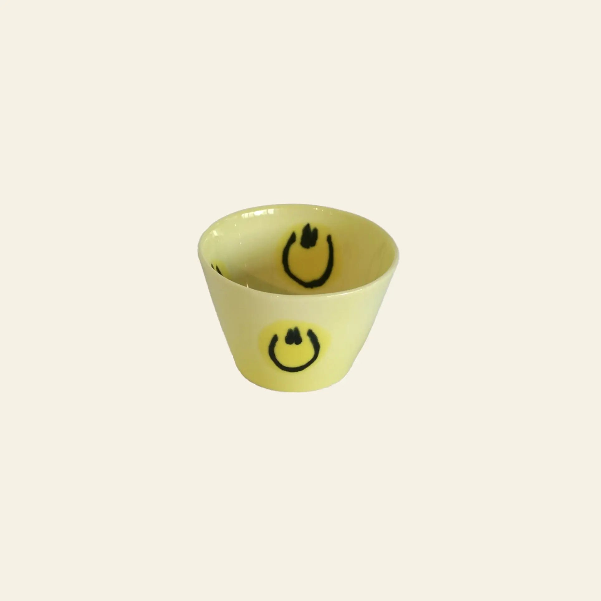 The Frizbee Ceramics Super Cup L by Frizbee Ceramics is a small yellow ceramic cup with black smiley face designs, adding artisanal charm and brightening any morning ritual.