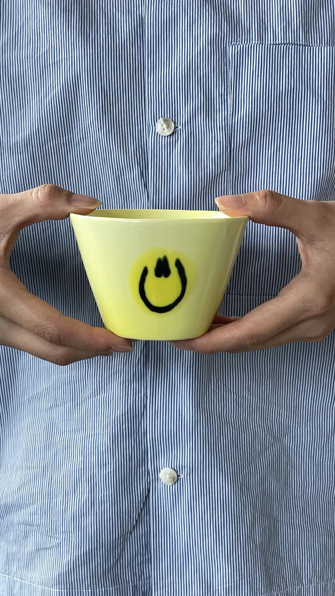 A person holds the Frizbee Ceramics Super Cup L, a yellow cup with a black smiley face, radiating artisanal charm—an ideal latte companion from Frizbee Ceramics.