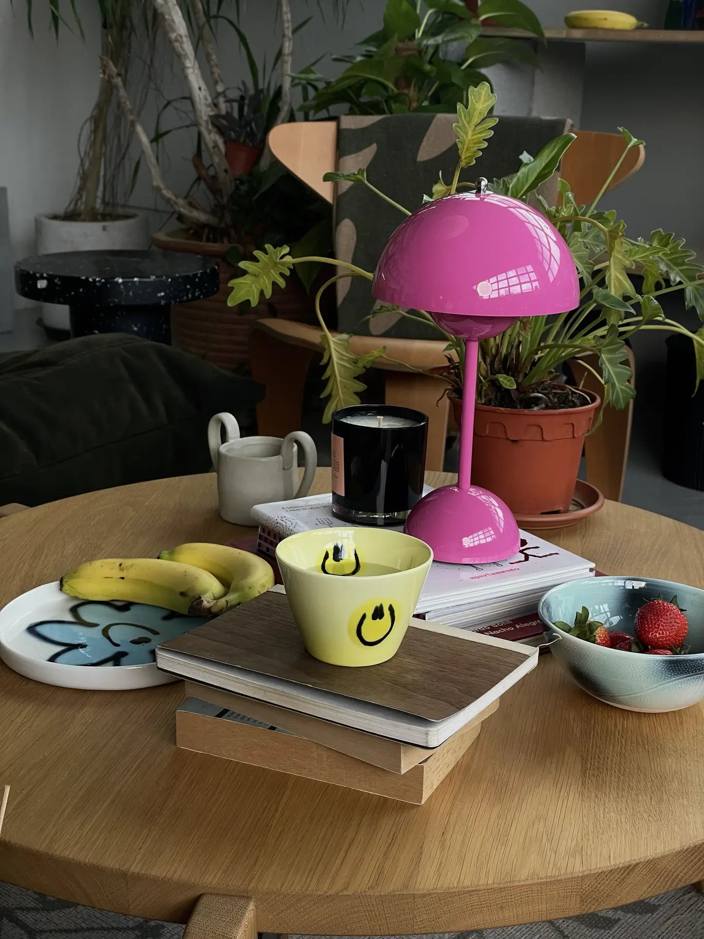A wooden table with books, a Frizbee Ceramics Super Cup L featuring black smiley faces, a yellow bowl, plate of bananas, strawberries, a pink lamp, black candle, and potted plants add artisanal charm to the cozy setting.