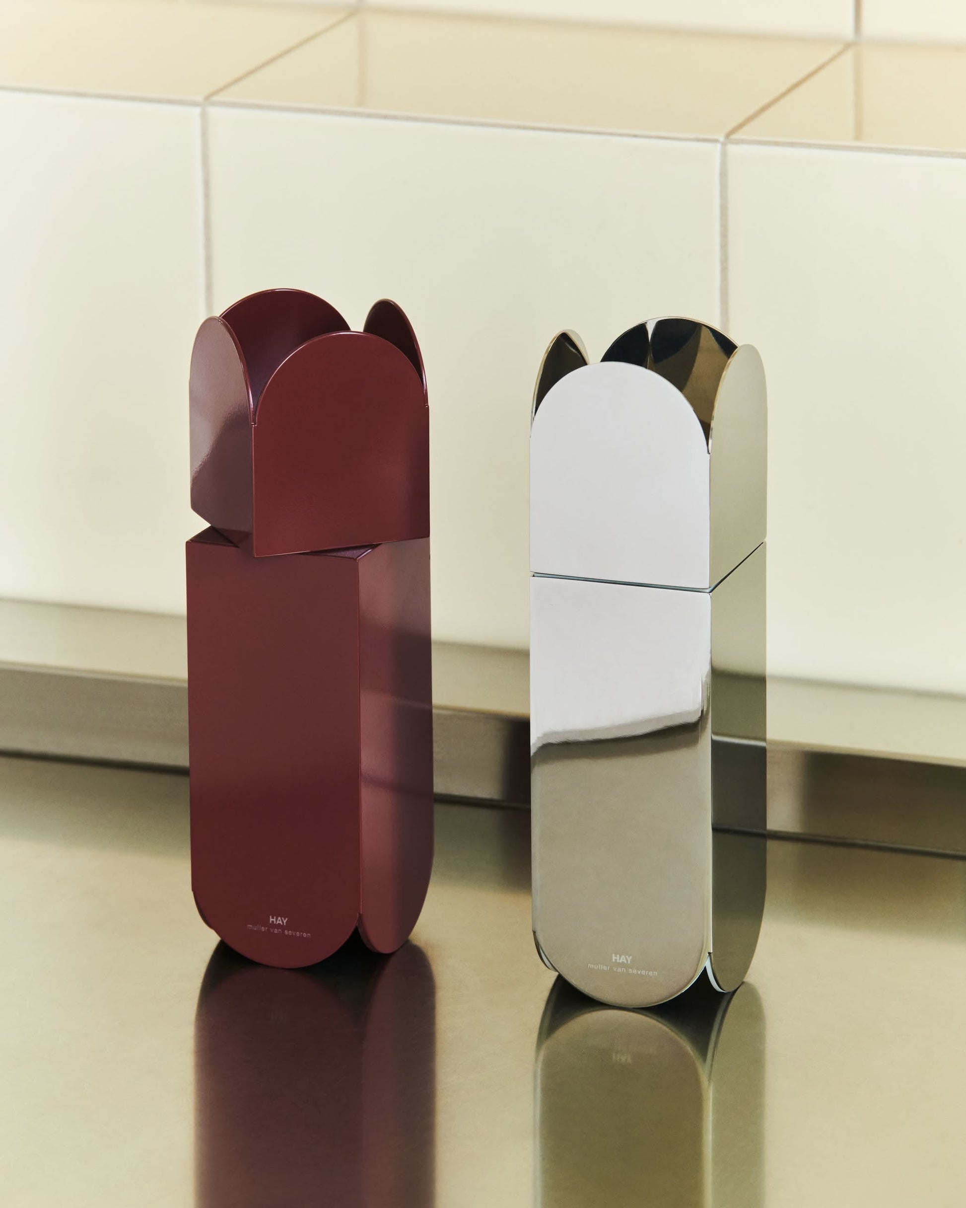 The HAY Arcs Salt & Pepper Grinders by HAY—one in glossy burgundy, one in shiny stainless steel—stand together on a metallic countertop. Both feature adjustable ceramic grinders for customizable seasoning.