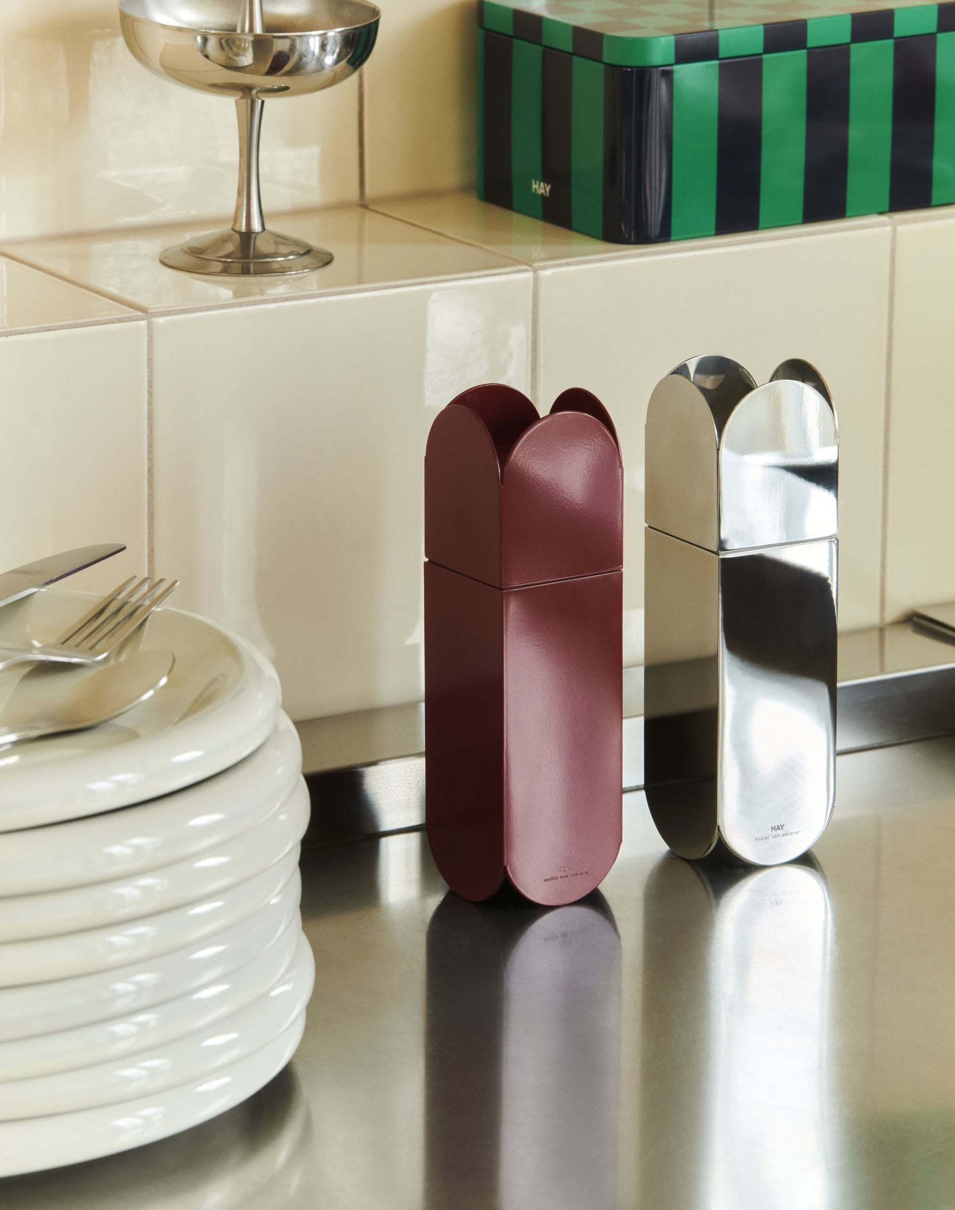 Two capsule-shaped HAY Arcs Salt & Pepper Grinders—one maroon, one stainless steel—by HAY stand upright on a kitchen counter near stacked white plates and cutlery. Each features an adjustable ceramic grinder.