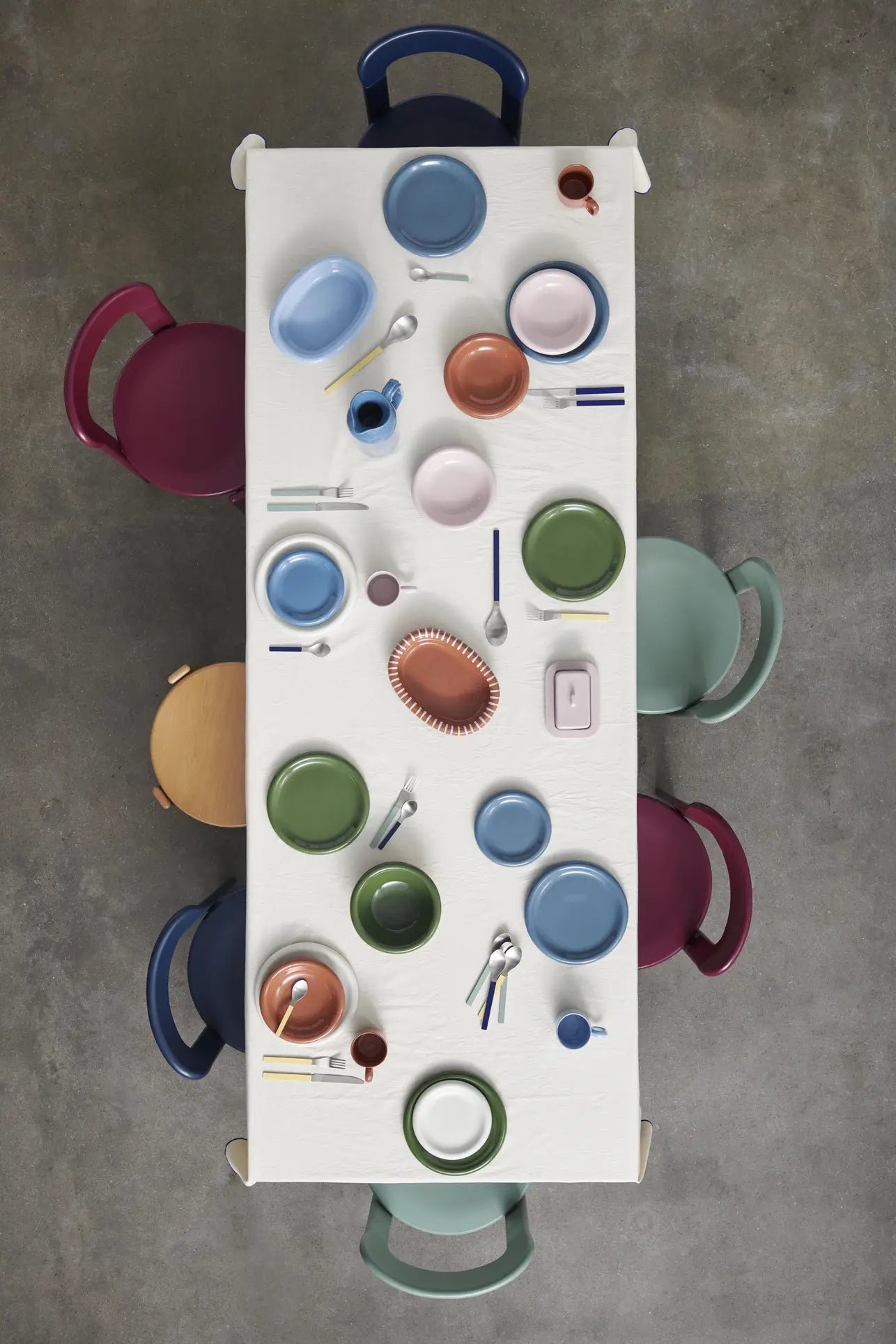 A rectangular table set for six showcases HAY Barro Bowl, Set of 2 by HAY with multicolored plates, bowls, cups, and Barro Bowl-inspired utensils on a white tablecloth; six colorful chairs surround it on a gray tiled floor.
