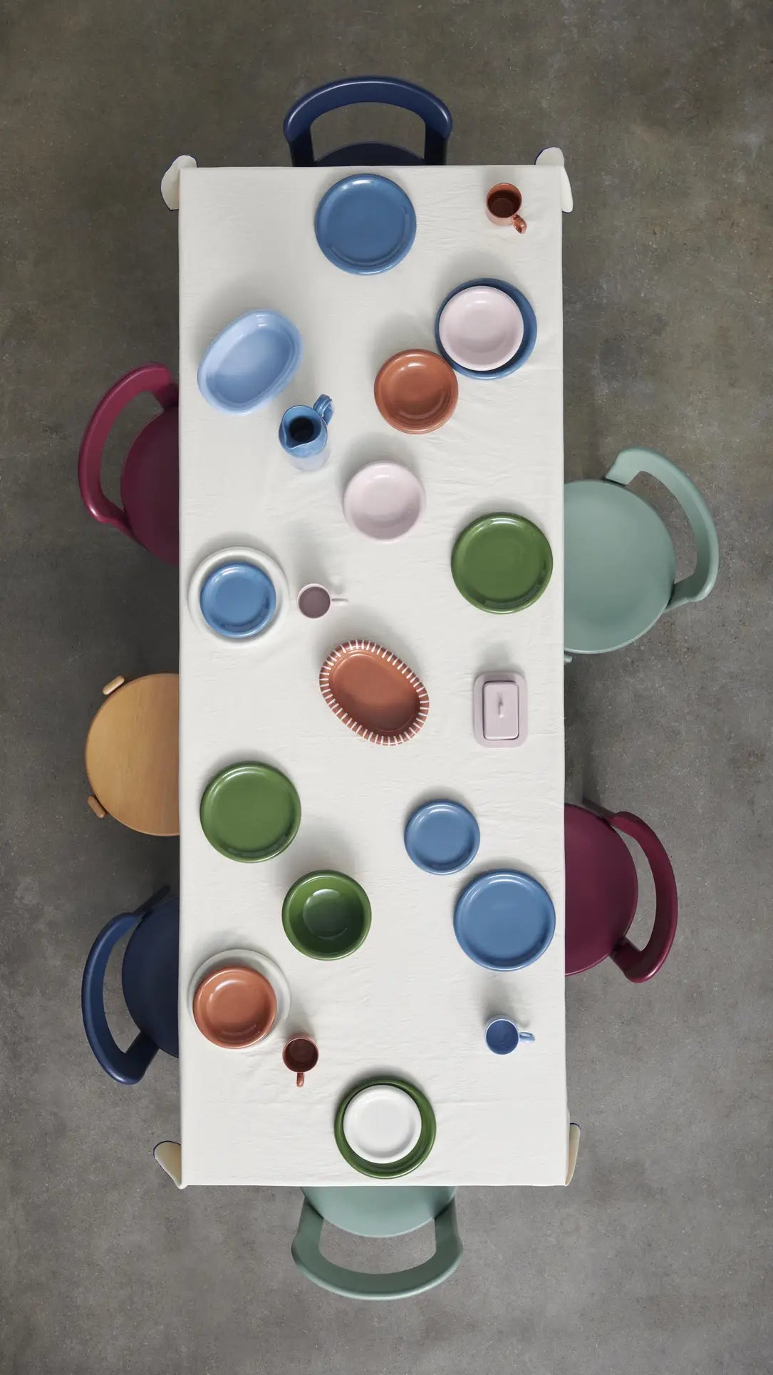 Overhead view of a rectangular table with a white tablecloth, colorful plates and cups, and HAY Barro Bowl, Set of 2 by HAY. Five multicolored chairs surround the table on a gray floor.