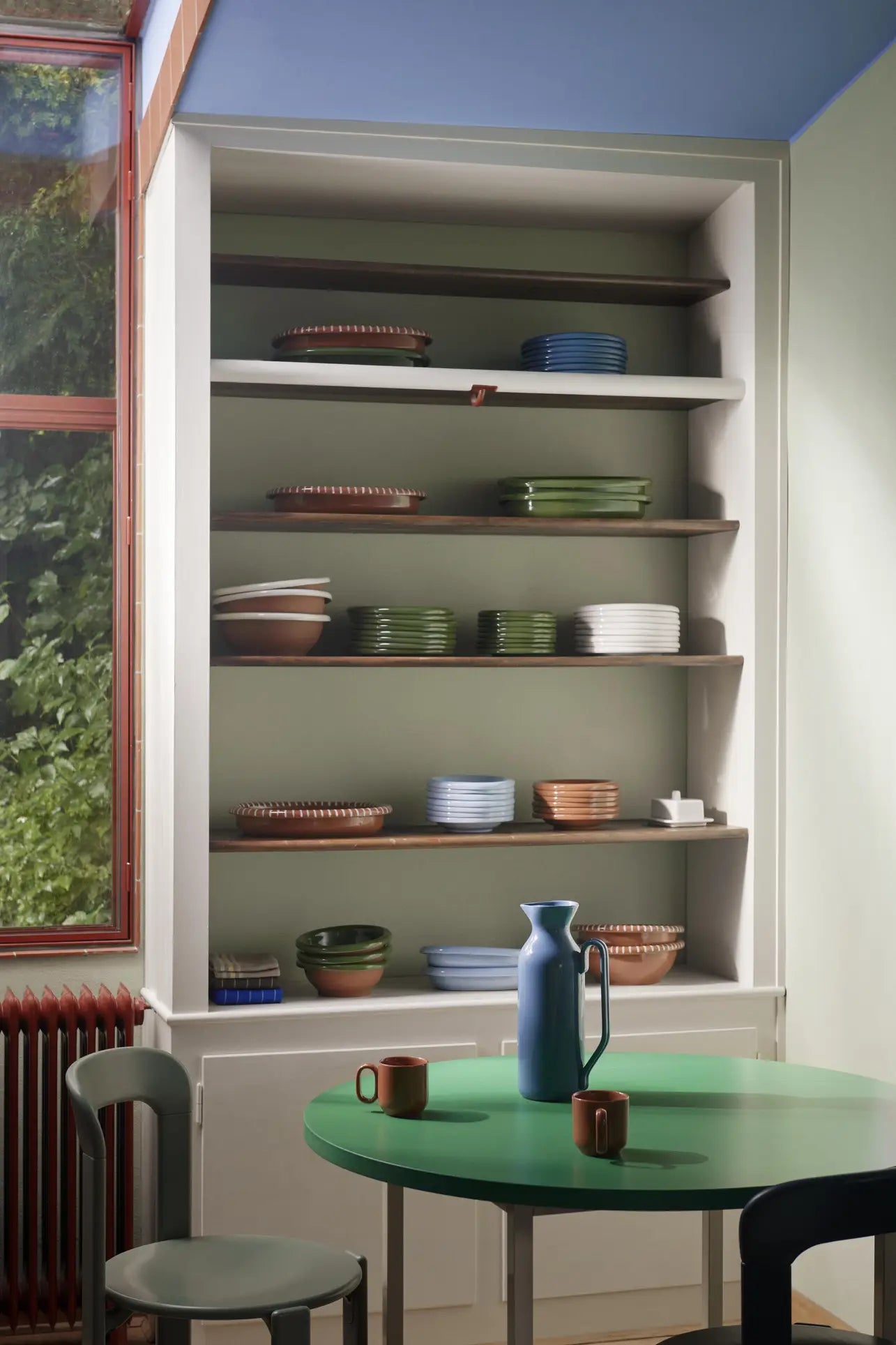 A built-in kitchen shelf displays neatly arranged plates and a HAY Barro Bowl, Set of 2 by HAY in earthy terracotta tones. Nearby, a round green table holds mugs and a blue jug, with greenery visible through the large window.