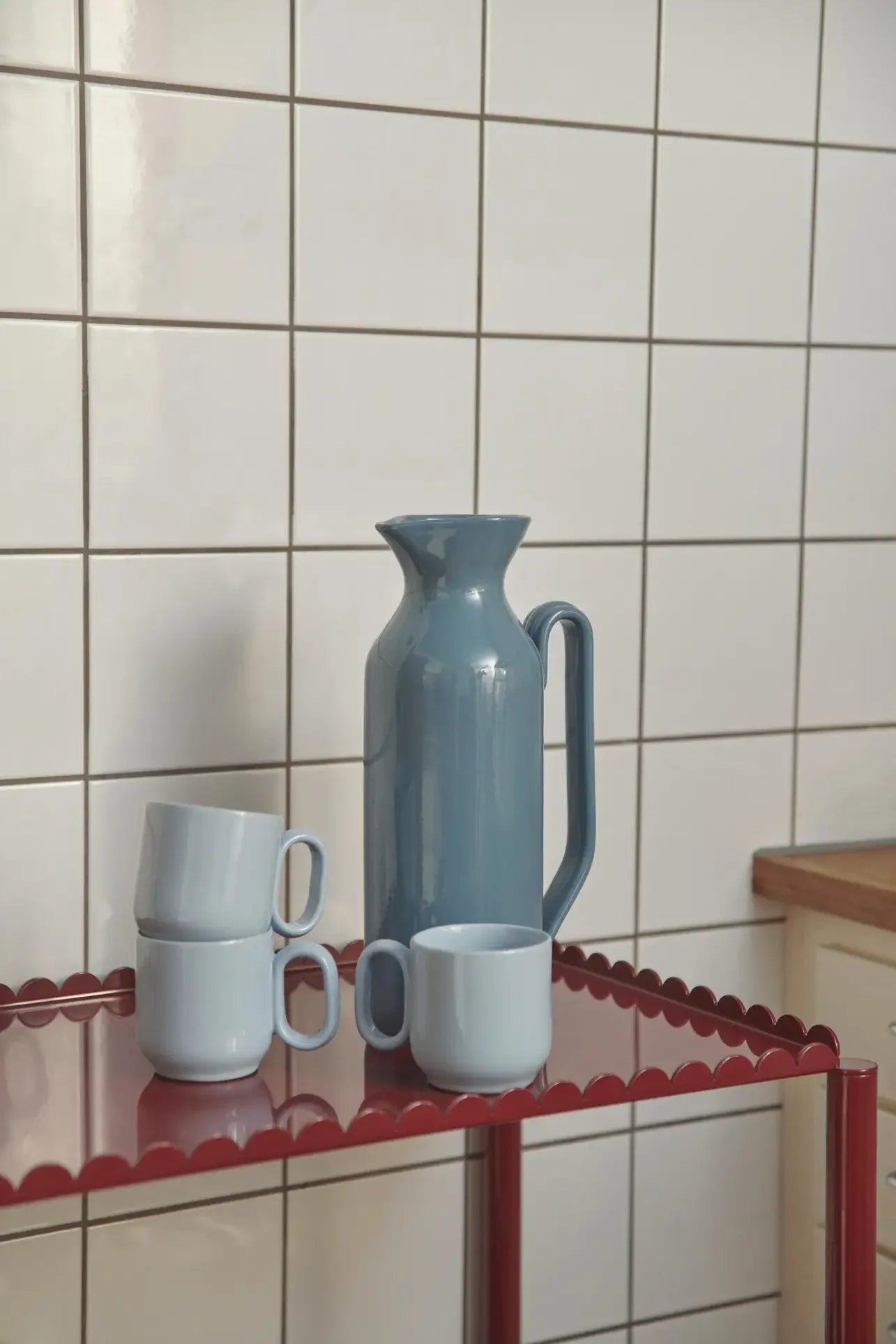 A tall blue ceramic pitcher and three stacked light blue mugs, similar to the HAY Barro Cup, Set of 2 by HAY, sit on a red metal shelf in front of a white tiled kitchen wall.