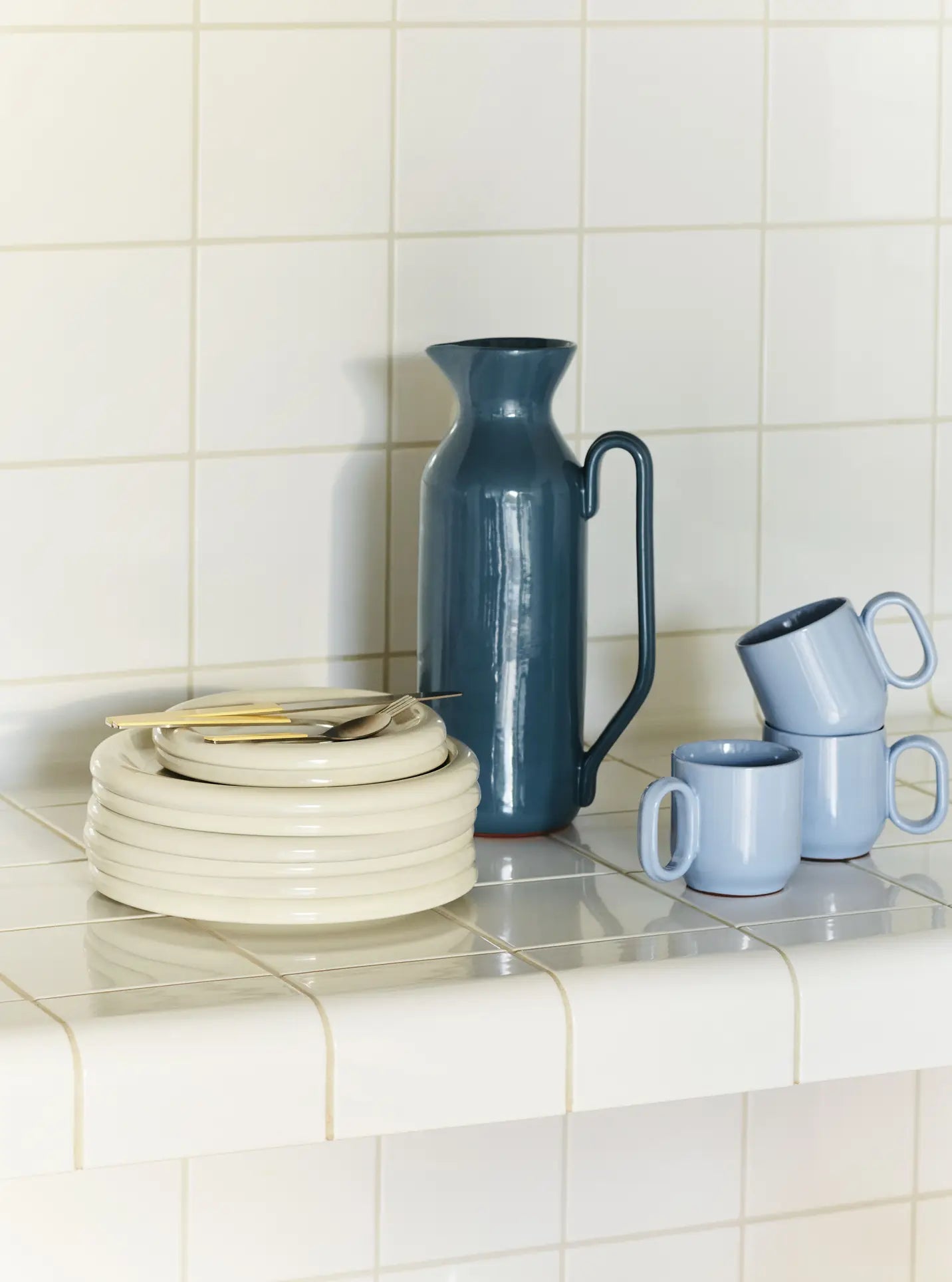 A stack of cream plates with cutlery, a tall blue pitcher, three light blue mugs, and the HAY Barro Cup by HAY (set of 2) are arranged on a white tiled countertop against a white tiled wall.