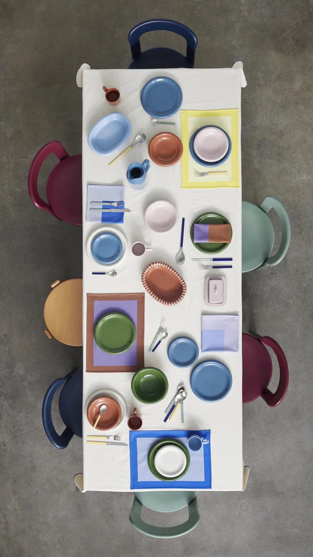 A rectangular table set for six features colorful terracotta HAY Barro Cups (Set of 2) and tableware, arranged on pastel placemats with six mismatched chairs, all set atop a gray floor.