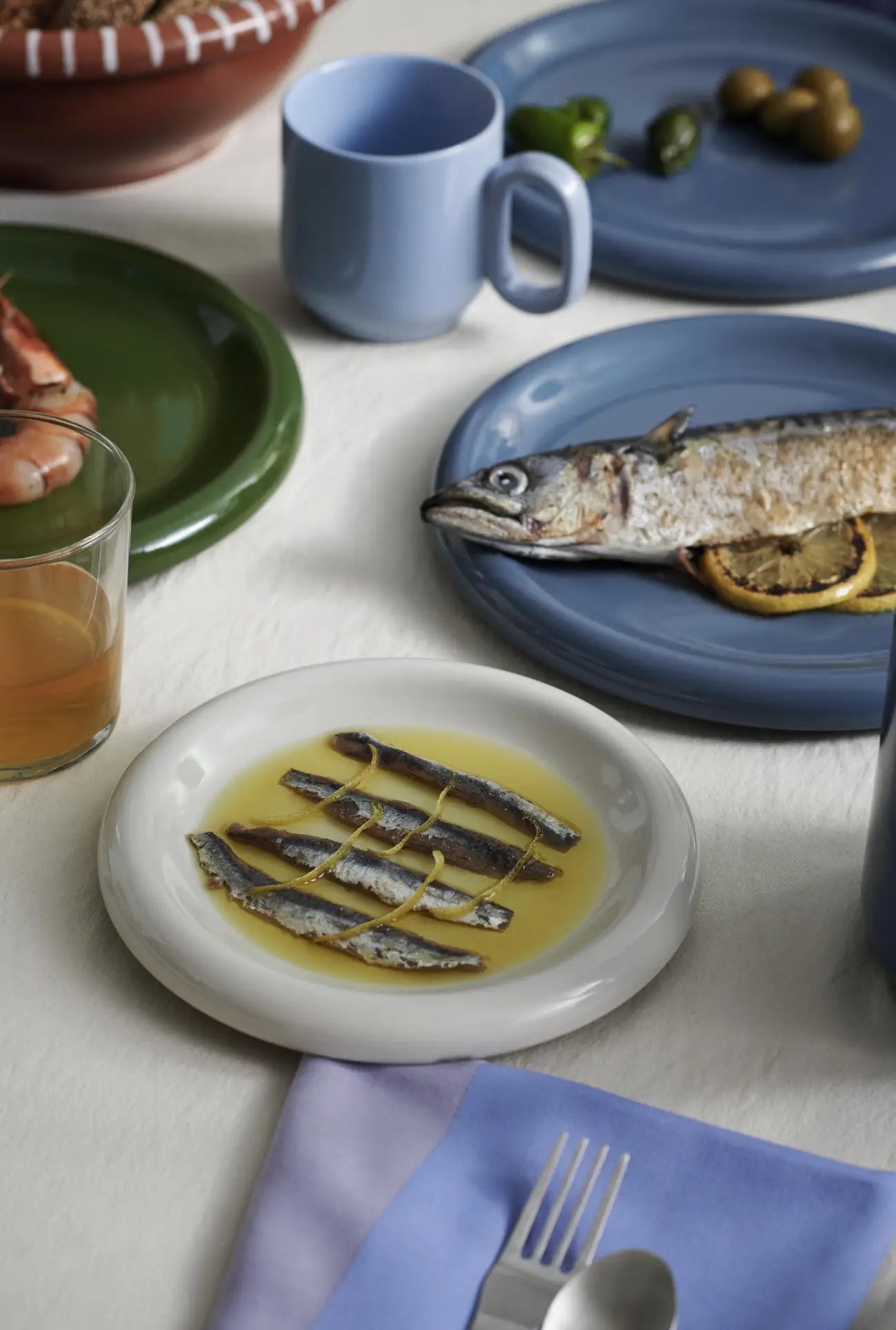 A table set with blue, green, and terracotta tableware features seafood, olives, and drinks. In the foreground, a HAY Barro Cup from the HAY Barro Cup Set of 2 sits beside a fork and spoon on a lavender napkin.