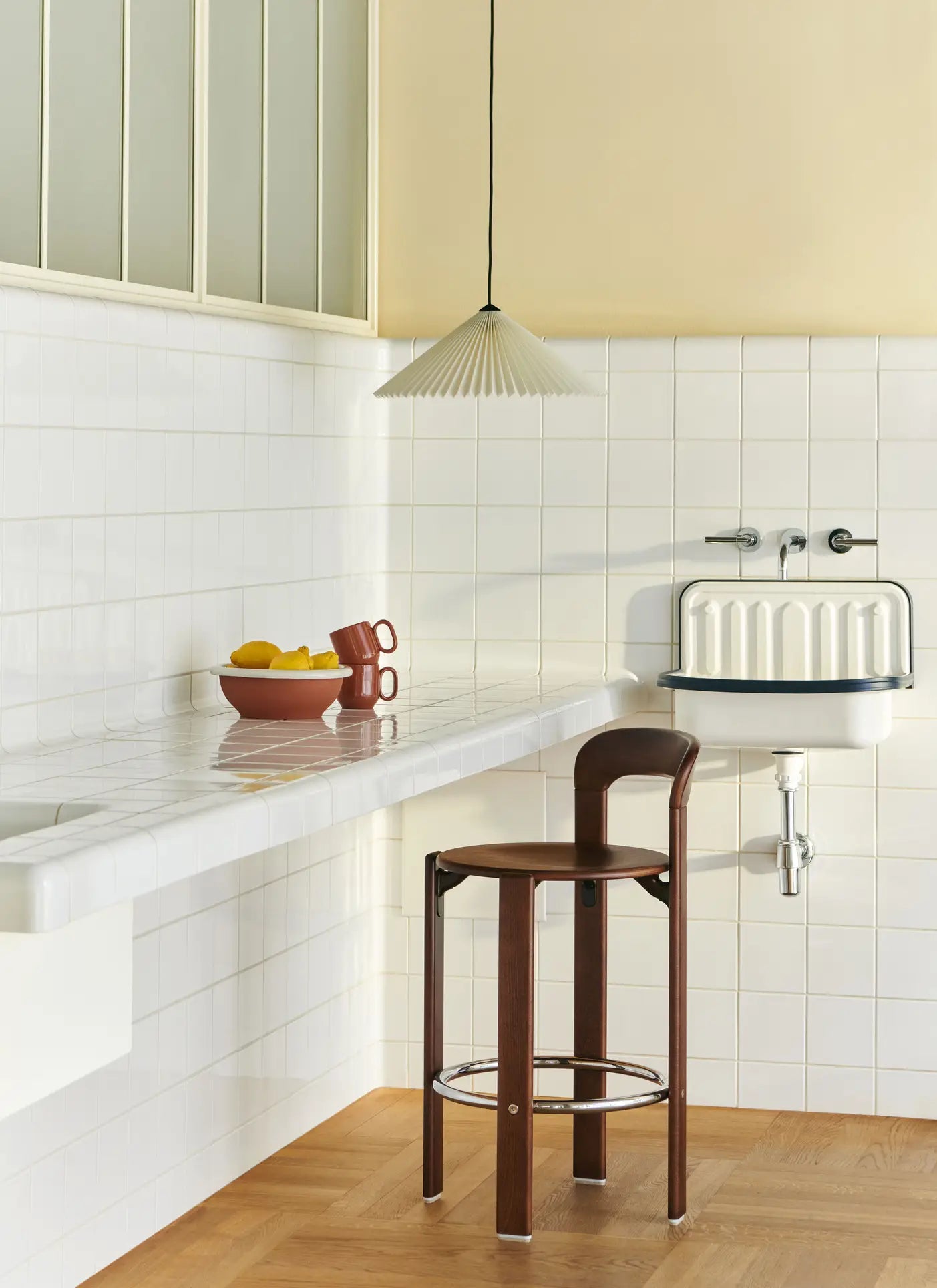 A minimalist kitchen with white tiled walls, a floating countertop holding lemons, terracotta tableware, the HAY Barro Cup Set of 2 by HAY, a brown wooden stool, vintage-style sink, and a pleated pendant light above.