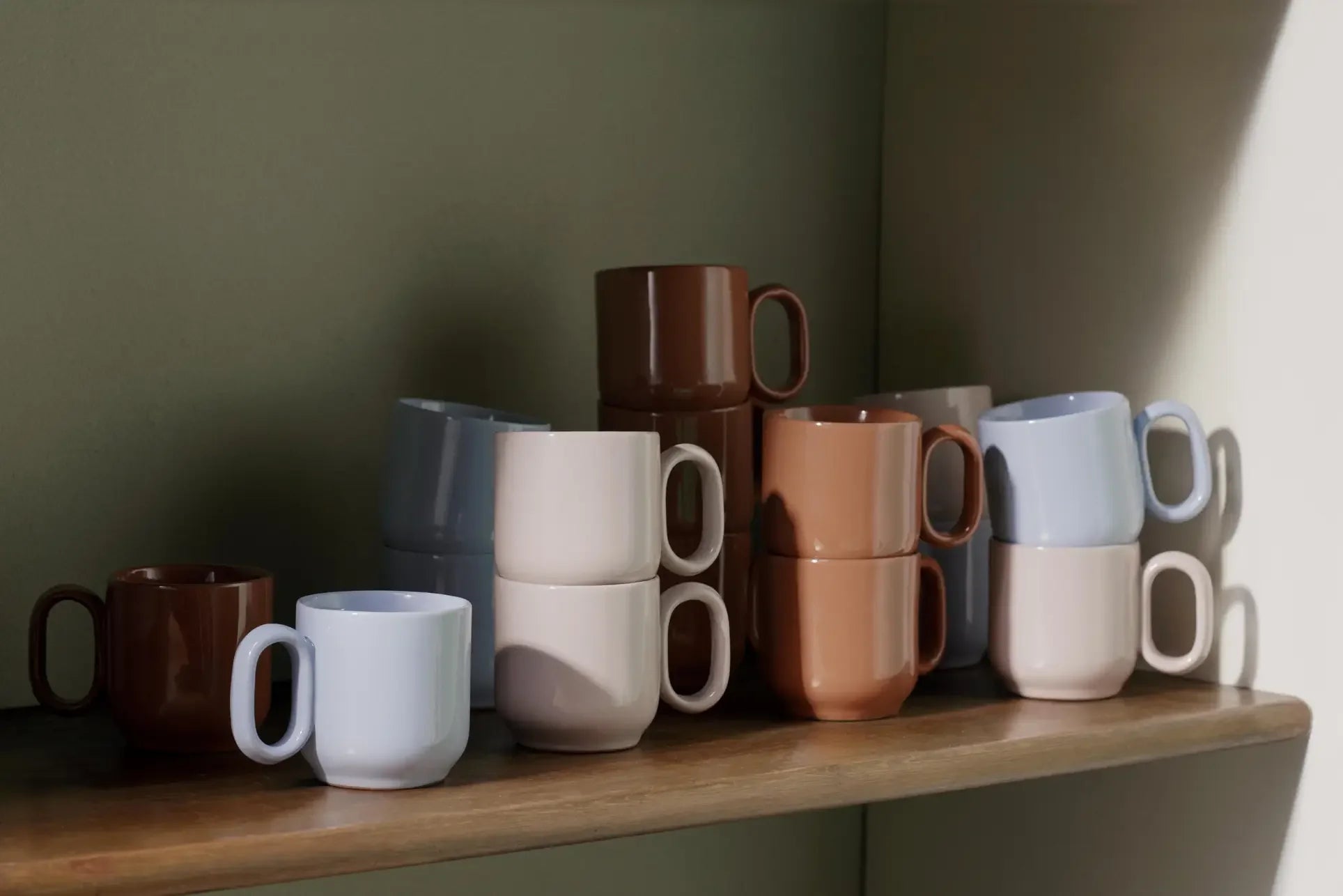 A set of HAY Barro Cups, designed by Rui Pereira, in neutral brown, beige, and light gray tones are stacked on a wooden shelf—showcasing the brand’s terracotta-inspired ceramic tableware style.