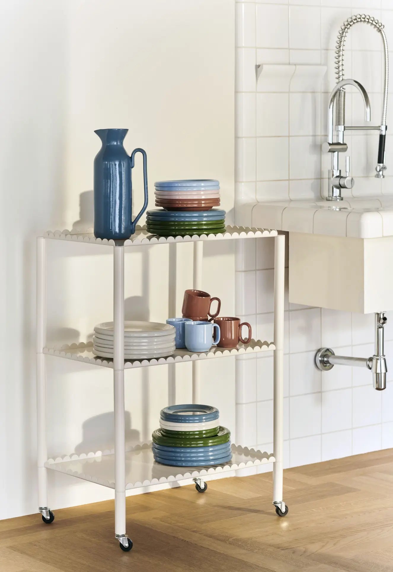 A cream-colored rolling cart with scalloped edges holds stacked plates, mugs, a blue pitcher, and a set of HAY Barro Cups by HAY, next to a white tiled sink in a kitchen with wooden flooring.