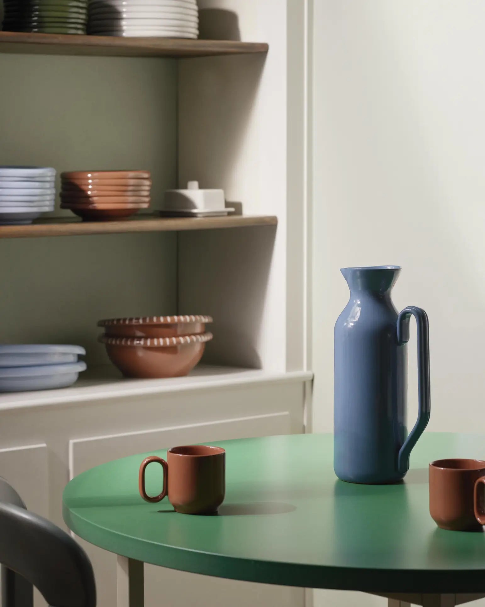 A green round table with a HAY Barro Cup set of 2 in brown and a tall blue ceramic pitcher. In the background, shelves showcase terracotta tableware in earthy tones, all within a softly lit, modern kitchen inspired by Rui Pereira.