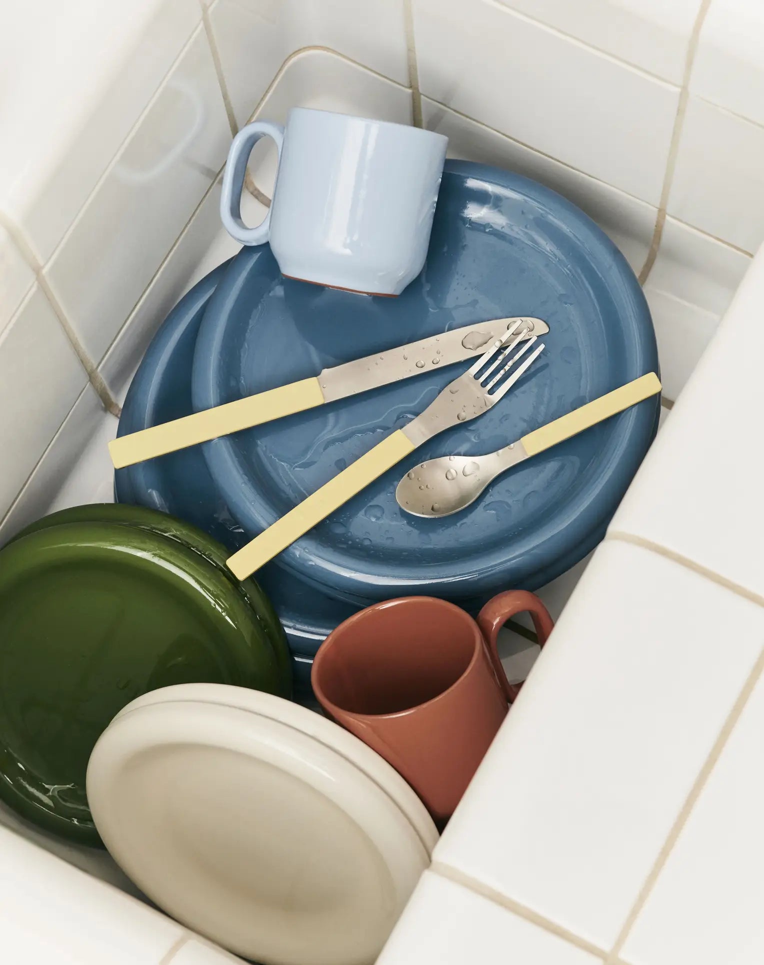 A kitchen sink with stacked dirty plates, cups, and cutlery. Some wet dishes include terracotta tableware inspired by the HAY Barro Cup, Set of 2 by HAY. The tiles around the sink are white.