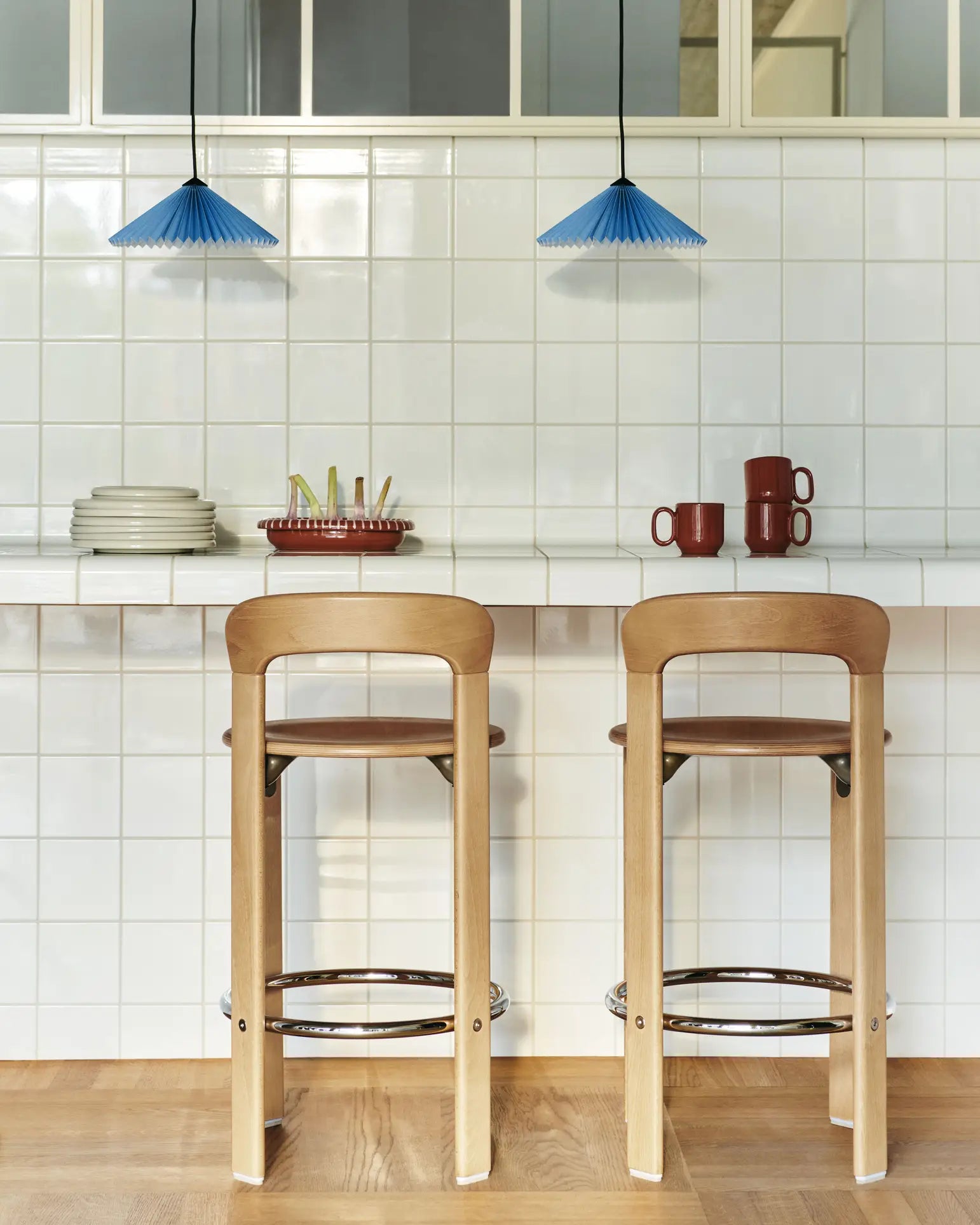 Two wooden bar stools sit at a white tiled counter with stacked plates, mugs, and a fruit tray, featuring the HAY Barro Cup Set of 2 by HAY. Overhead, two blue pendant lights complete this clean and minimalistic setting.
