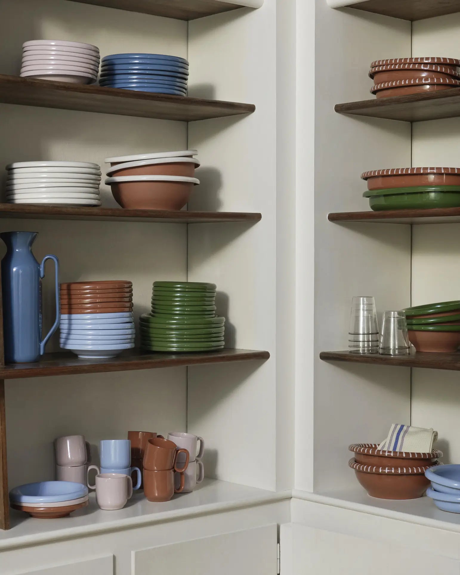 Shelves in a corner kitchen cabinet display neatly stacked HAY Barro Cups, earthy-toned terracotta tableware, a blue pitcher, folded striped towels, and other pieces inspired by Portuguese designer Rui Pereira.