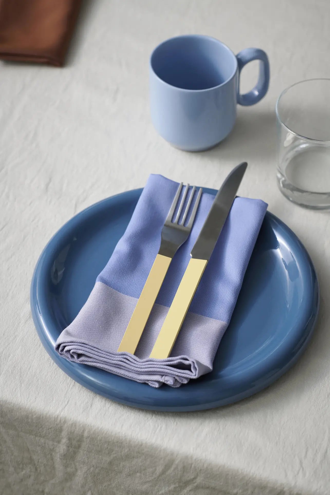 A HAY Barro Plate 24cm (set of 2) by HAY, with a folded lavender napkin and gold-handled fork and knife, rests on a light tablecloth. Nearby are a blue mug, clear glass, and brown napkin from the tableware collection.