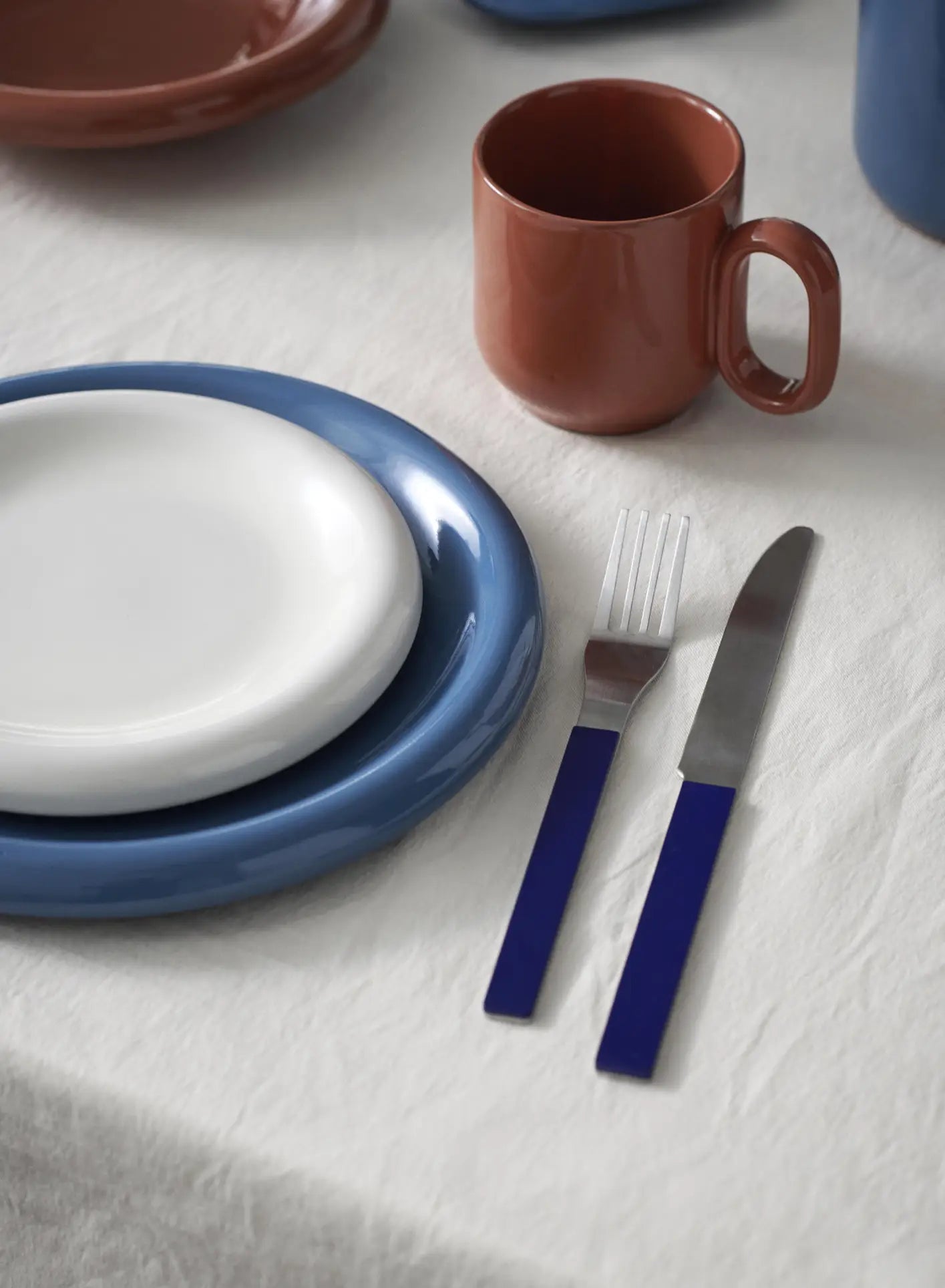 A neatly set table with a white cloth features a HAY Barro Plate 24cm, Set of 2 by HAY stacked on a blue plate, a brown terracotta mug, and blue-handled cutlery—showcasing an elegant tableware collection.