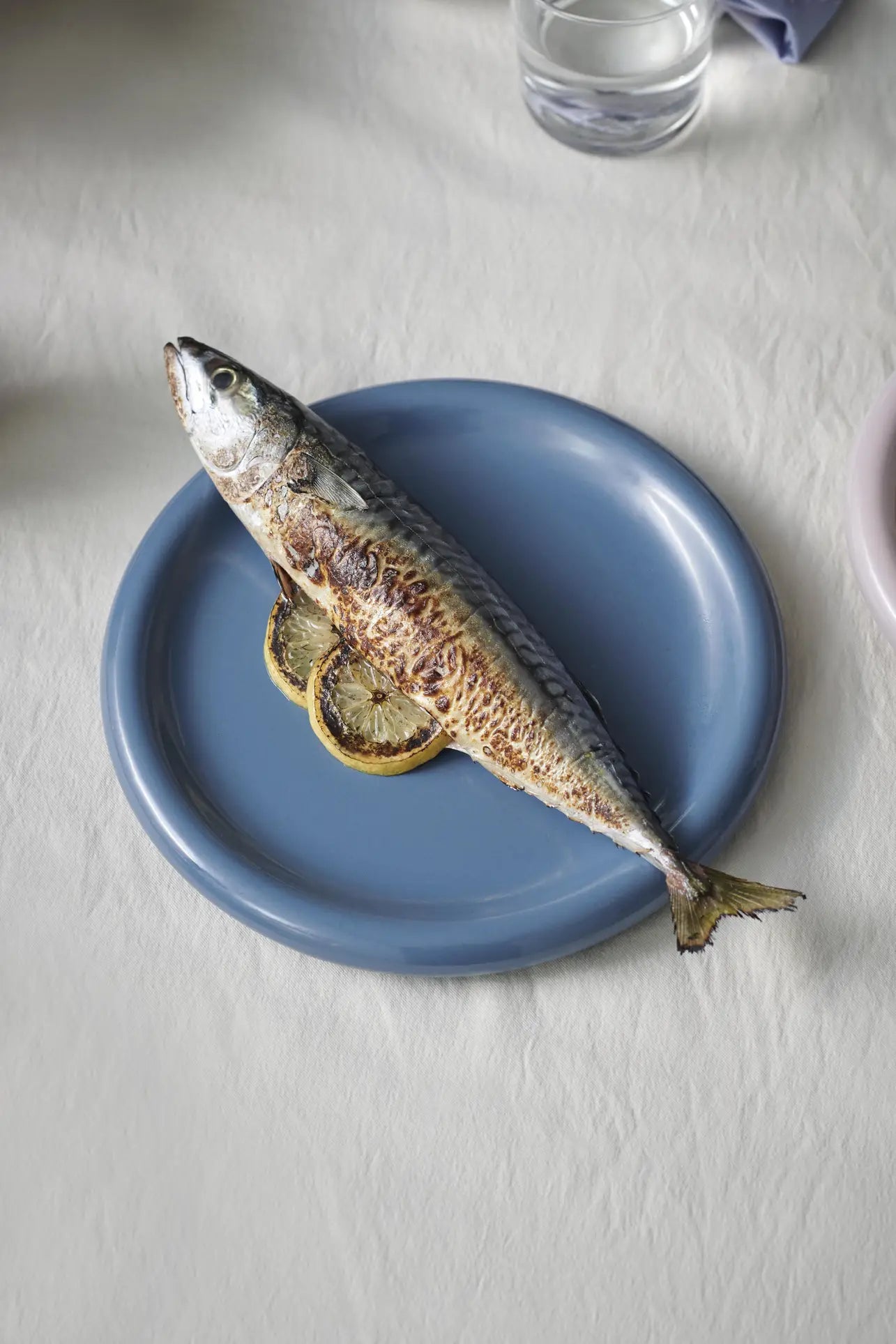 A whole grilled fish sits on lemon slices atop the HAY Barro Plate 24cm from HAY, part of a set of 2, arranged on a white tablecloth with a glass of water nearby.