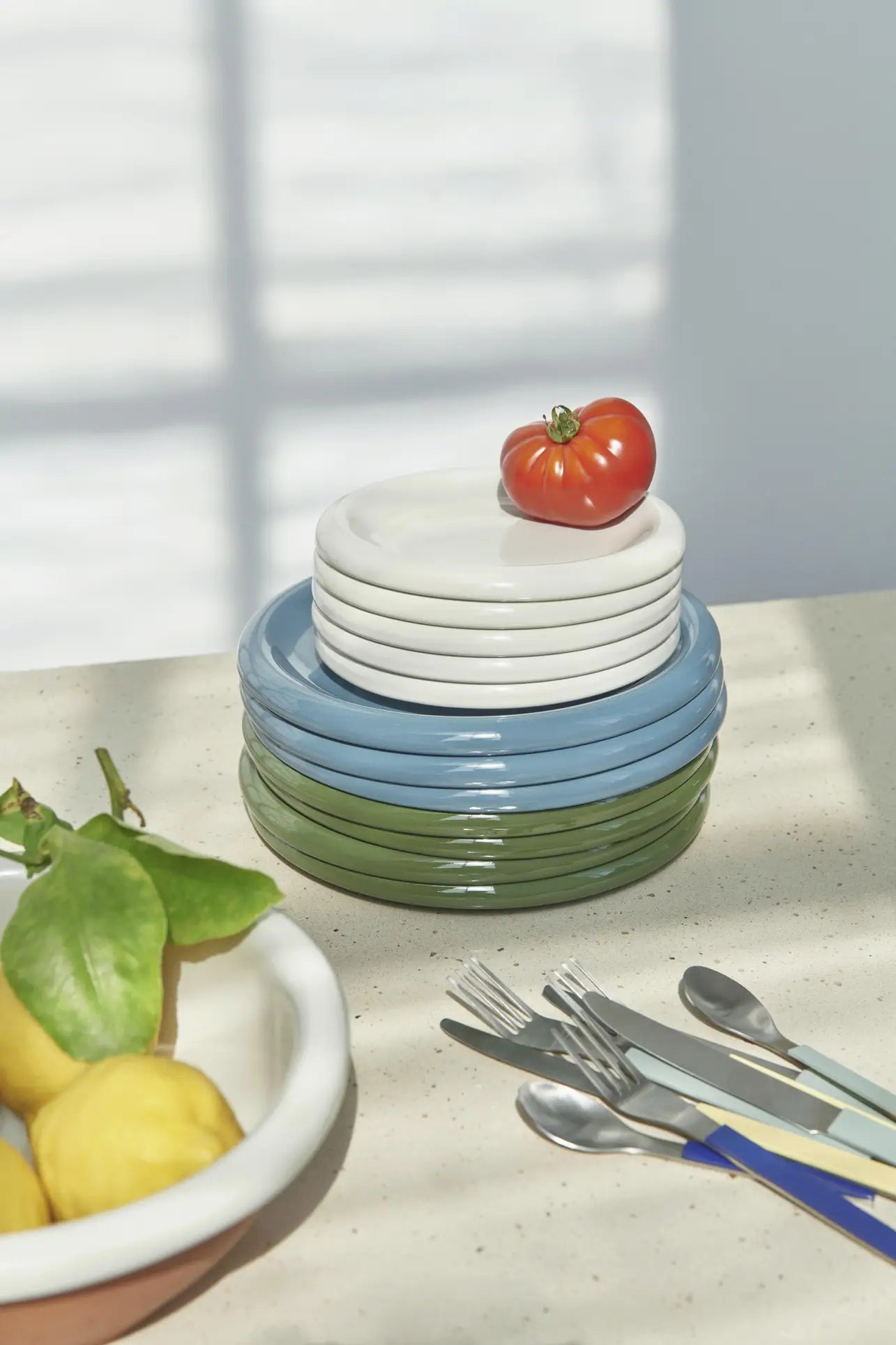 A stack of HAY Barro Plate 24cm, Set of 2 in white, blue, and green sits on a light countertop with a red tomato on top. Nearby are lemons with green leaves in a bowl and colorful-handled cutlery, all bathed in soft sunlight.