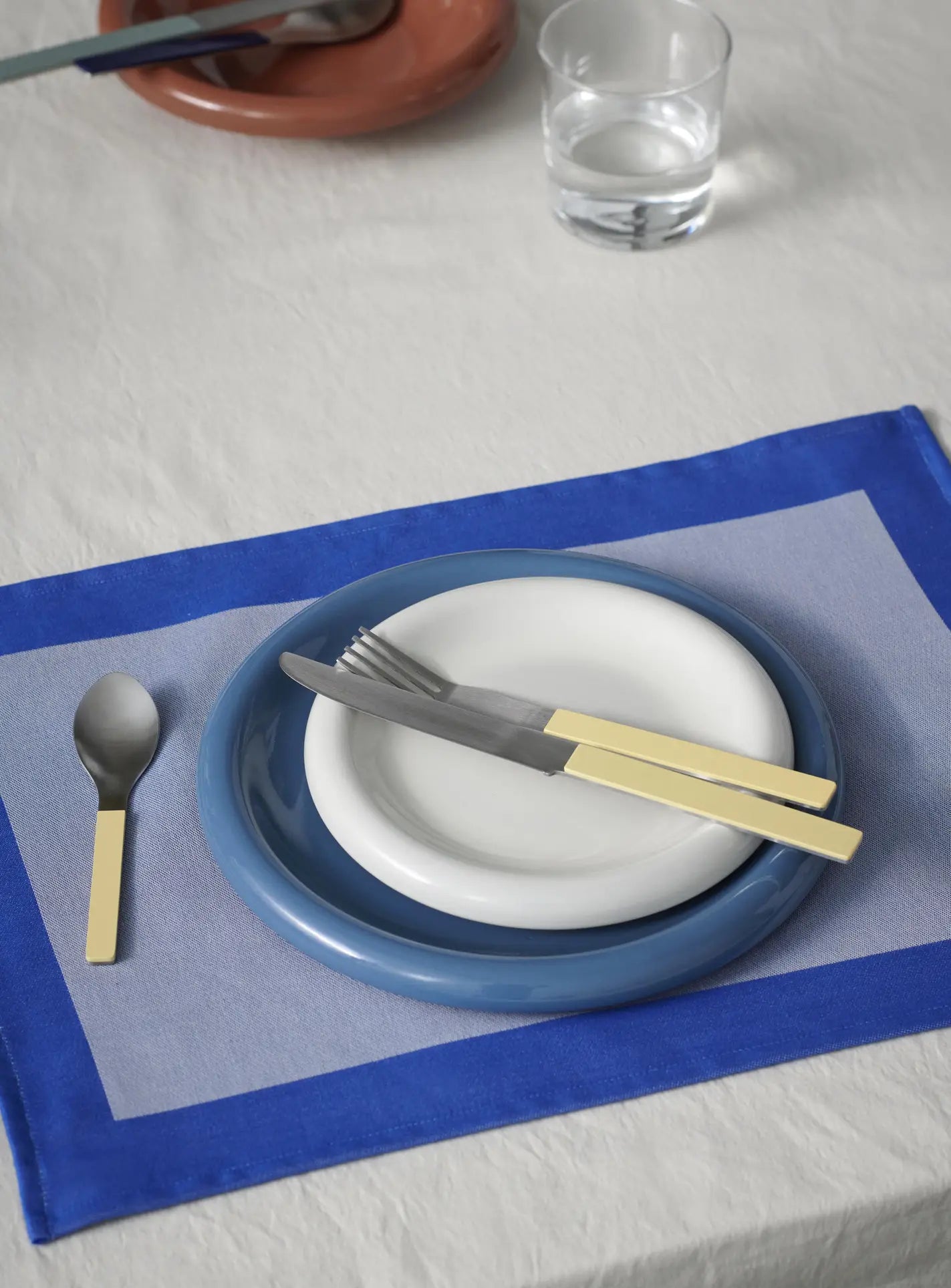 A table setting with a blue-bordered placemat, a stacked HAY Barro Plate 24cm and white plate, yellow-handled fork and knife diagonally on the plates, spoon to the left, and a glass of water in the background.